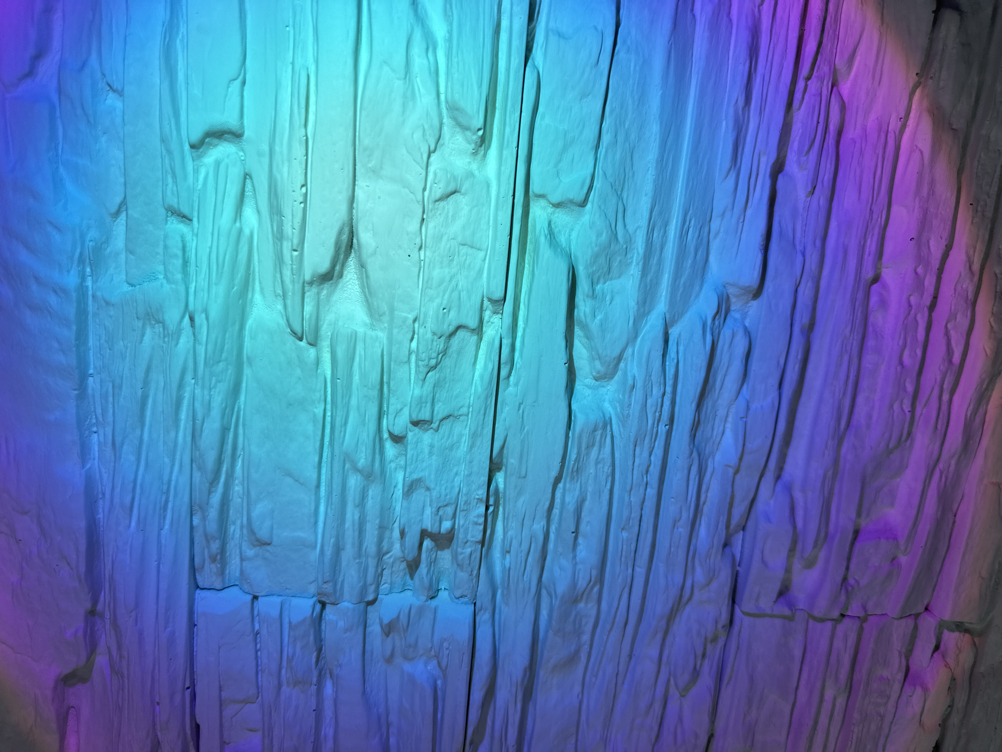 The image depicts a textured wall, possibly made of stone or a similar material, illuminated with a vibrant, multicolored light gradient. The lighting shifts from blue on the left to teal in the center and transitions to purple on the right, creating a dramatic visual effect. The wall is constructed of vertical, slightly uneven blocks or panels, giving it a somewhat rugged appearance. There are no people or other identifiable subjects present in the image. The location and city are Unknown, Unknown.