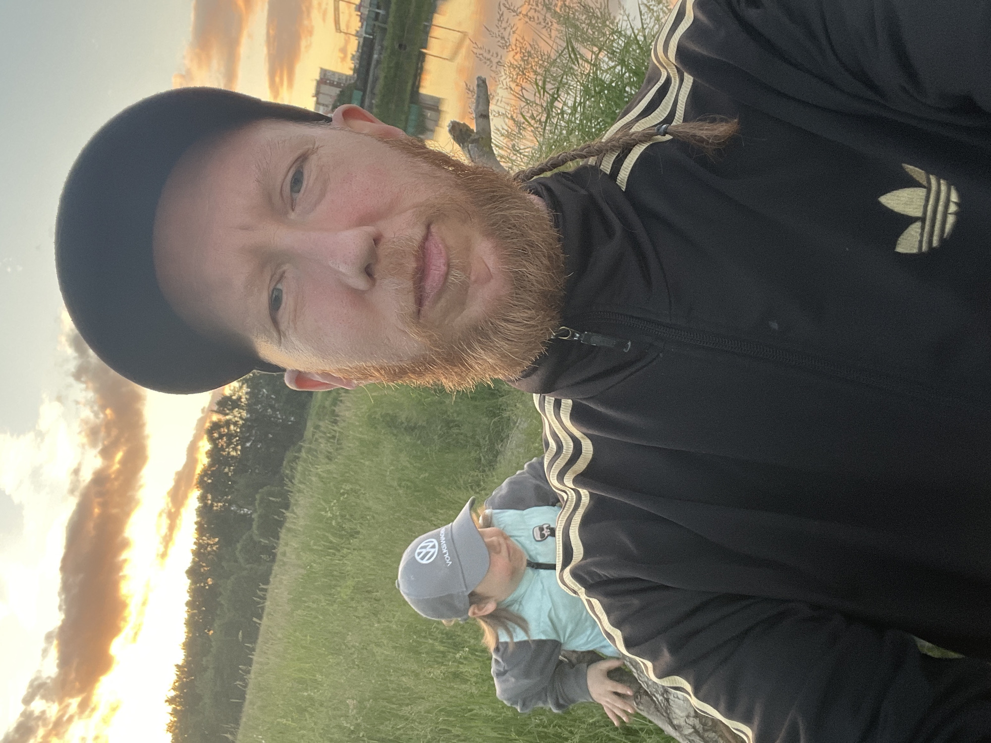 A selfie taken by a man with a red beard and a black baseball cap, wearing a black Adidas tracksuit with gold stripes, shows him in the foreground. Behind him, a child wearing a grey baseball cap with the Volkswagen logo and a teal vest over a gray jacket is seen sitting on a log. The background features a grassy hill, a line of trees, a body of water reflecting the sunset with colorful clouds, and buildings in the distance. The image appears to be taken at sunset, likely in a park or natural area near the city. Given the description of a city with canals, a historical port, and a famous sunset spot, this could be the area around Krestovsky Island in Saint Petersburg, Russia, which offers similar views of water, greenery, and cityscapes.