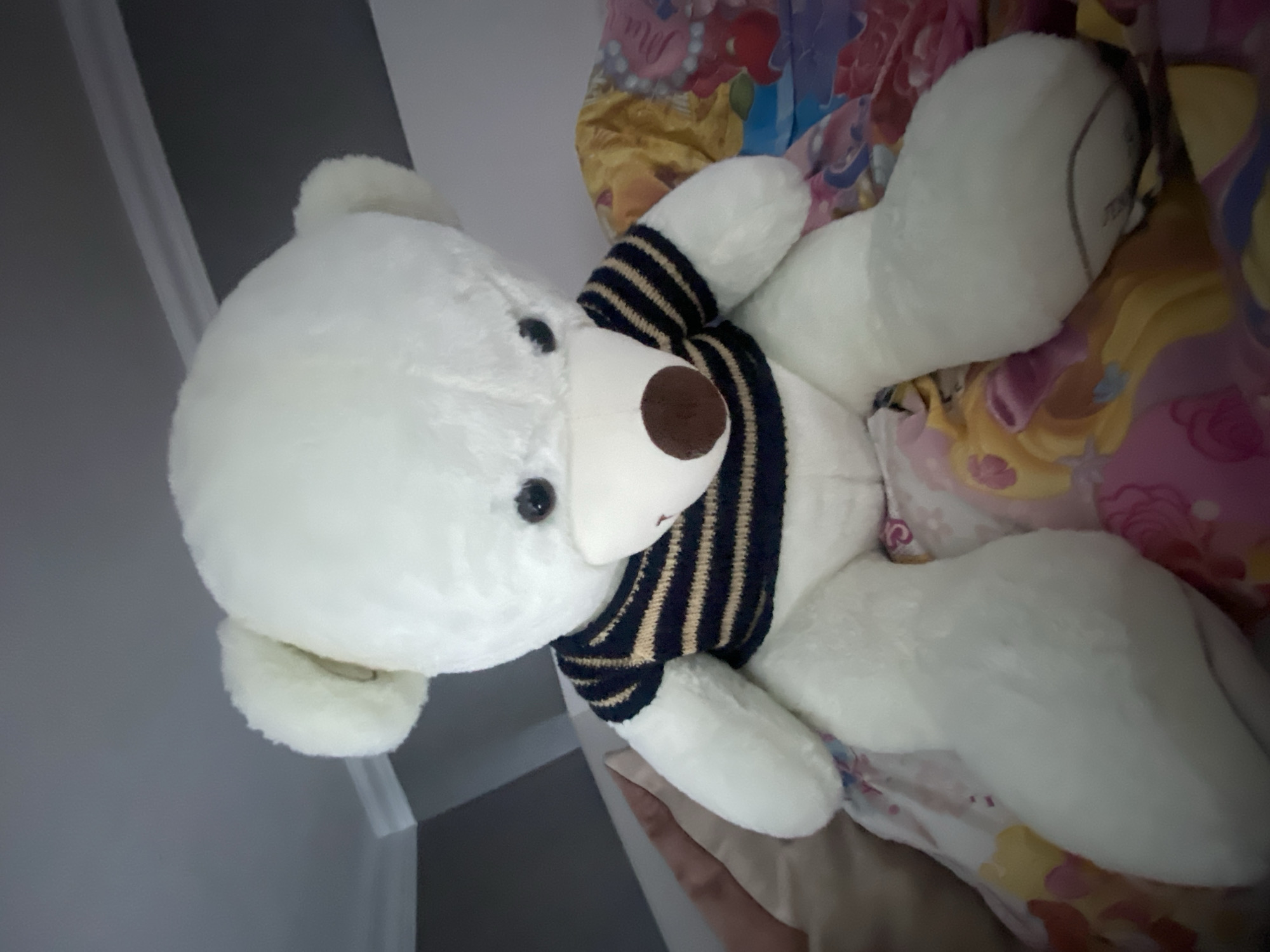 A white teddy bear wearing a striped sweater sits on a colorful bedspread. The teddy bear is positioned facing forward, with its head slightly bowed. Its fur is fluffy, and it has dark eyes and a brown nose. The sweater has horizontal stripes of navy blue and tan. The bedspread features a vibrant pattern with various colors and possibly cartoon characters. The background appears to be a room with a gray wall and white trim. The image seems to be taken indoors, likely in a residential setting. There is no indication of the city of Rybnitsa, Moldova, in the visual information provided.