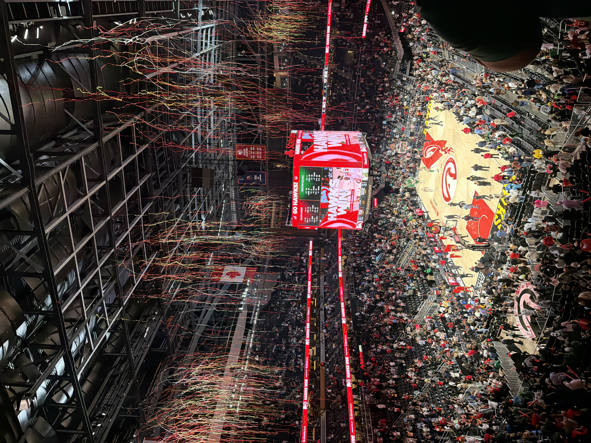 Here is a description of the image:
The image depicts a basketball game at State Farm Arena in Atlanta, Georgia, United States. The arena is filled with spectators, and celebratory confetti has been released. A large video board hangs over the court, displaying the game score and graphics for the Atlanta Hawks, the home team. The Hawks have apparently won the game. There are red columns on either side of the arena. A Canadian flag can be seen at the left, also. The environment is festive and energetic, indicative of a sports victory.