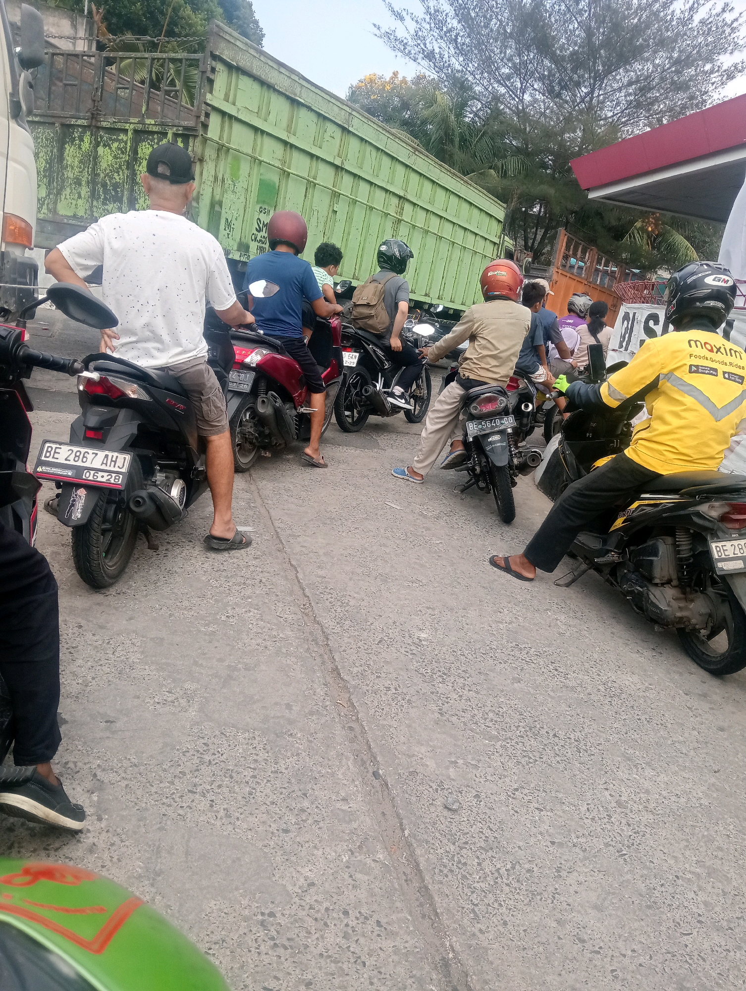 In the city of Bandar Lampung, Indonesia, a group of people on motorcycles are waiting in traffic. The scene is set on a road with a grey, pebbled surface. In the background, a large green truck is parked, with its side doors open. To the right of the truck, a red and white awning, possibly from a gas station or convenience store, is visible. The sky above is clear and bright, suggesting daytime.
Several individuals are present, all on motorcycles. One person in the foreground, wearing a white t-shirt with black speckles and a black baseball cap, is seated on a dark-colored motorcycle. Their legs are bare, and they are wearing grey cargo shorts and dark flip-flops. Another person, facing away from the camera and wearing a blue t-shirt and red helmet, is on a red motorcycle. Further back, a person in a tan jacket and light-colored pants is on a motorcycle. To their right, another person in a purple shirt is visible. On the far right, a person in a bright yellow jacket with a "Maxim" logo on it is seated on a dark motorcycle, wearing a black helmet. Their hands are covered in light green gloves. Other individuals are partially visible in the background, also on motorcycles. The motorcycles are of various colors, including black, red, and dark grey. One motorcycle in the foreground has a license plate that reads "BE 2867 AHJ". The overall impression is a moment of stillness in a potentially busy urban environment, with people waiting for traffic to move.