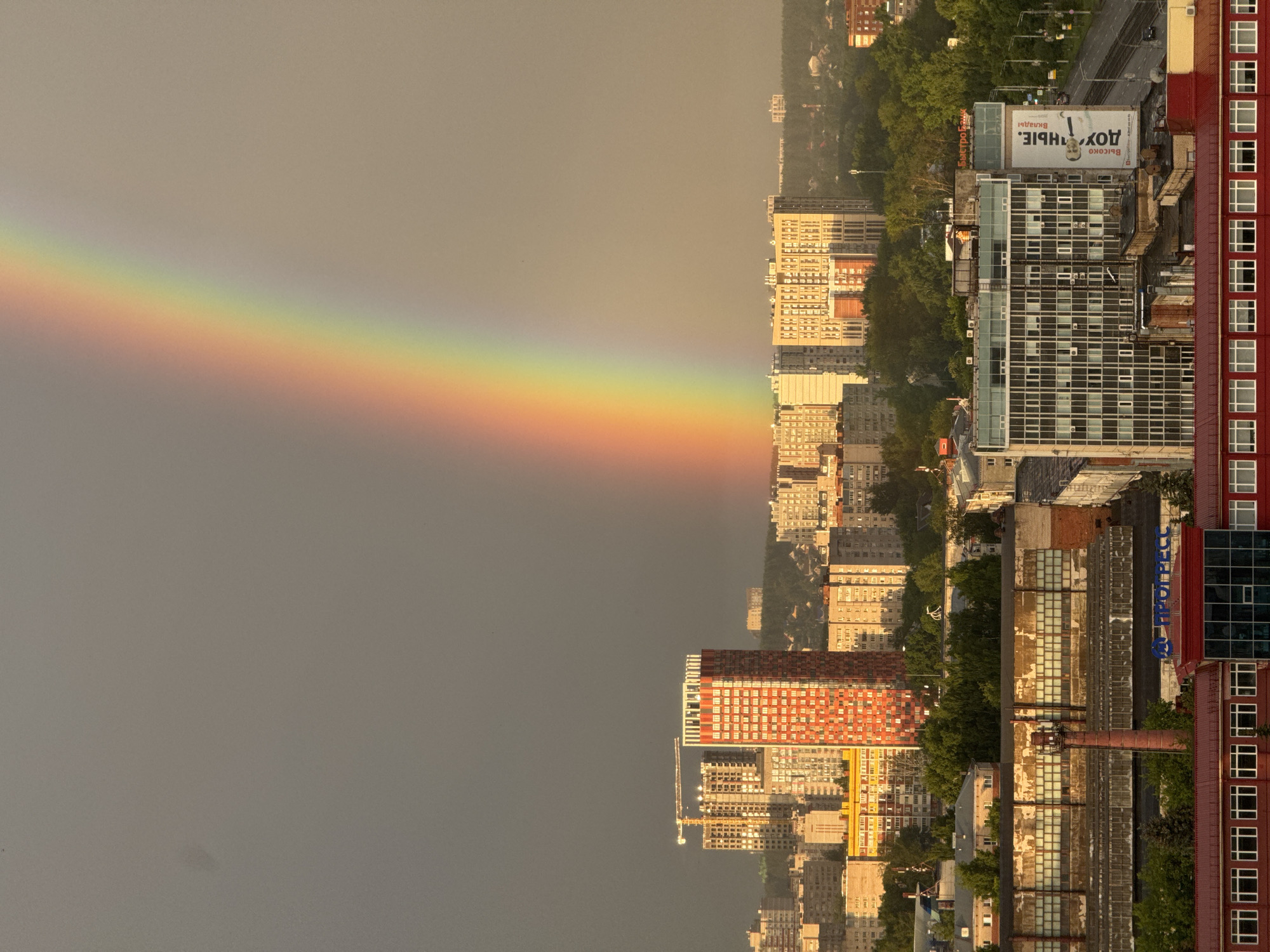 The image captures a cityscape in Izhevsk, Russia, under a dusky sky, illuminated by the warm glow of the setting sun. A vibrant rainbow arcs across the sky, its colors vivid against the muted tones of the clouds. In the foreground, an industrial building with a red roof and numerous windows is visible, with a tall brick chimney to its left. To the right of this building, a modern, multi-story office building stands, featuring a facade of glass and white panels, and a large banner advertising "ДОУЧНЫЕ. ВКЛАДЫ" with a portrait of a man. Beyond these structures, rows of residential buildings and a tall, orange-colored skyscraper dominate the middle ground, some under construction, indicated by a crane. The background is filled with a dense forest and more buildings, fading into the hazy horizon. The overall scene suggests a post-rain atmospheric condition, common in the region.