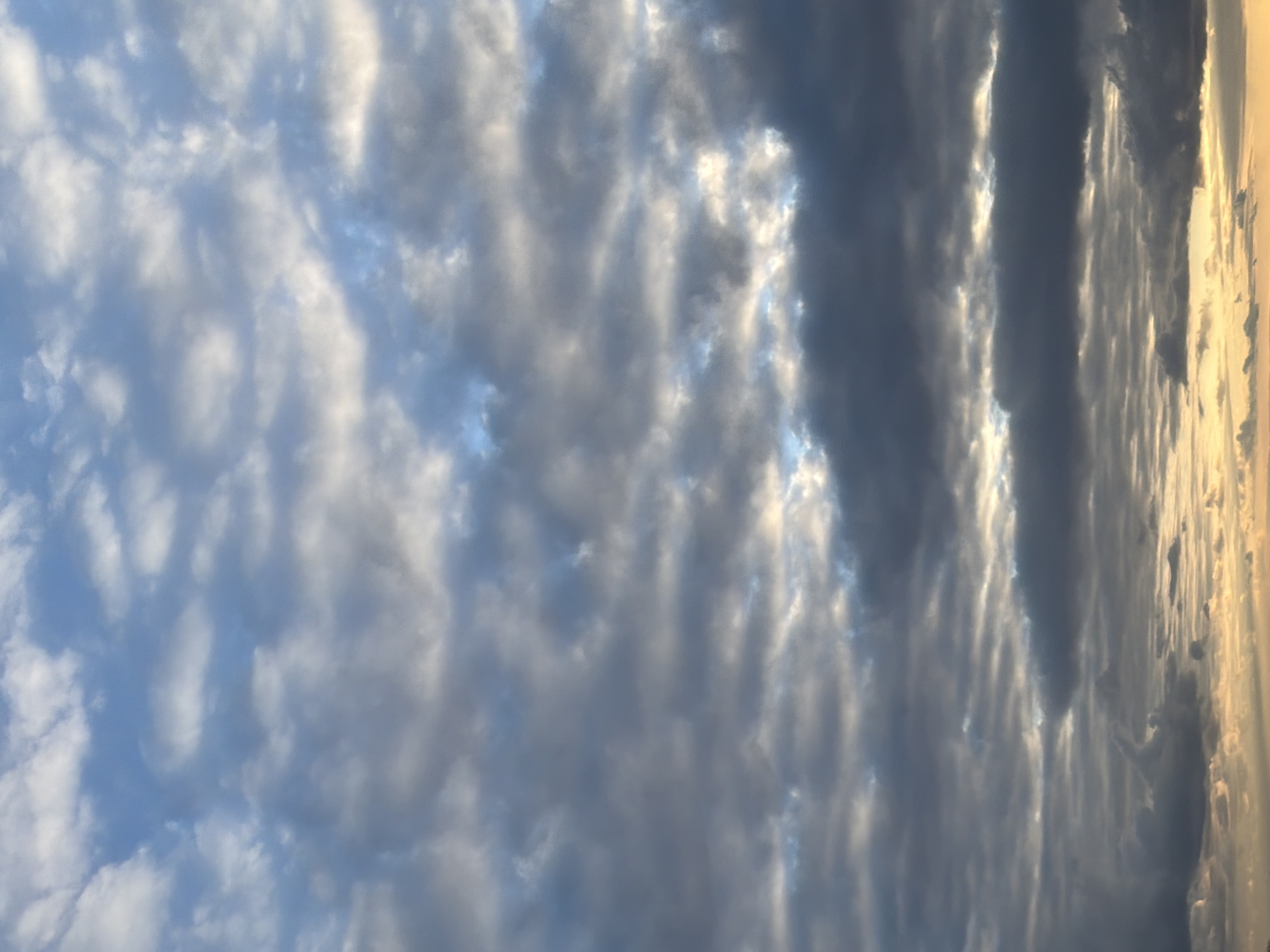 The image presents a dramatic and expansive view of a cloudy sky, dominated by various cloud formations and atmospheric lighting. The left side of the frame features a lighter blue sky dotted with soft, white to light-grey clouds, which appear somewhat broken and textured, allowing patches of the clear blue sky to show through.
Moving towards the center and right, the cloud cover becomes significantly denser and darker, appearing in shades of deep grey and blue-grey. Bright, intense streaks of golden and brilliant white light pierce through and behind these darker clouds, creating a stark and beautiful contrast. This strong illumination suggests the sun is positioned towards the far right of the scene, indicating either a sunrise or sunset, as the light appears to be originating from a low angle. The golden glow highlights the edges of the clouds, giving them a luminous quality.
The interplay of light and shadow across the clouds creates depth and a sense of volume within the sky. There are no visible people, objects, or identifiable geographical features, making the sky and its changing conditions the sole subject of the photograph. The overall scene conveys a powerful and dynamic natural phenomenon, capturing the beauty of clouds illuminated by a low sun. No text is visible in the media file.