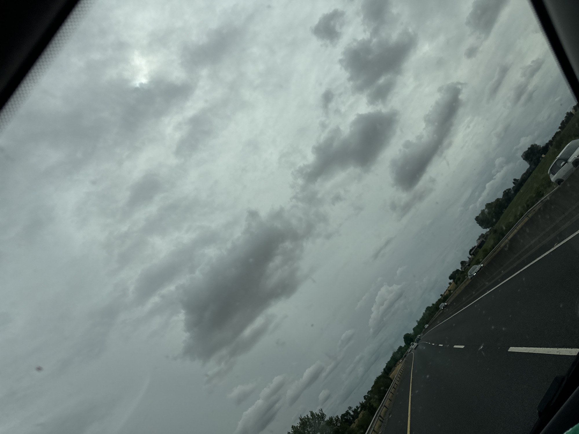 The image shows a view from a car on a highway, likely in or near Pill Belan, Ireland, given the cloudy sky and lush green landscape. The highway is paved and has clear white lane markings. Several cars are visible on the road, suggesting moderate traffic. The sky is filled with heavy, dark clouds, indicating overcast or possibly rainy weather. Trees and fields line the sides of the highway. The perspective is from the driver's side of the vehicle, looking forward and slightly to the right.