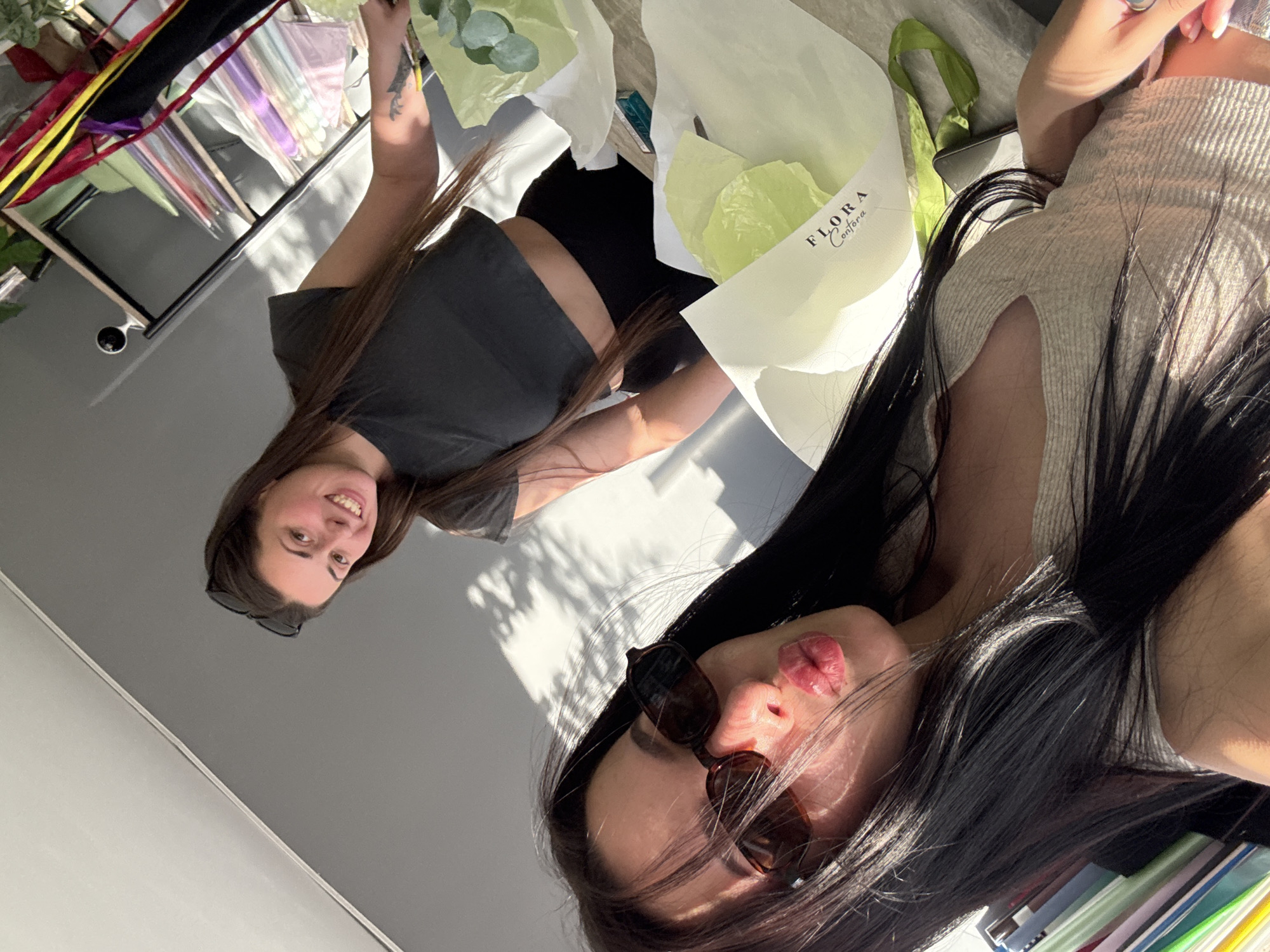 Two young women are captured in a selfie-style photograph. The woman in the foreground, with long dark hair, is wearing sunglasses and a light-colored, ribbed tank top, and is puckering her lips. She is positioned in the lower left quadrant of the image. Behind her and to the right, a second woman with long brown hair, wearing a dark, cropped t-shirt and black pants, smiles at the camera. She has sunglasses perched on her head. They appear to be in an indoor setting, possibly a workspace, with shelves filled with colorful materials and rolls of paper visible in the background to the right. A table in the middle ground holds wrapped bouquets, one with a label that reads "FLORA Confora." The lighting suggests natural sunlight casting shadows on the wall behind the women.