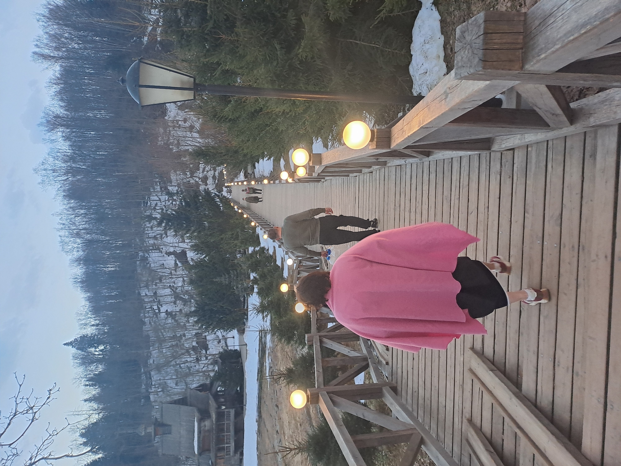 The image shows a downward-sloping wooden pedestrian bridge in what appears to be Vzglyadnevo, Russia. Several people are walking on the bridge. One individual, closest to the camera, is wearing a pink garment. Another person, farther down the bridge, is wearing green and black clothing. The bridge is lined with evenly-spaced, spherical lamps. The surrounding environment consists of trees, some bare and some evergreen, as well as areas with snow cover. A building is visible at the base of the bridge. A tall street lamp stands at the bridge's highest point.
