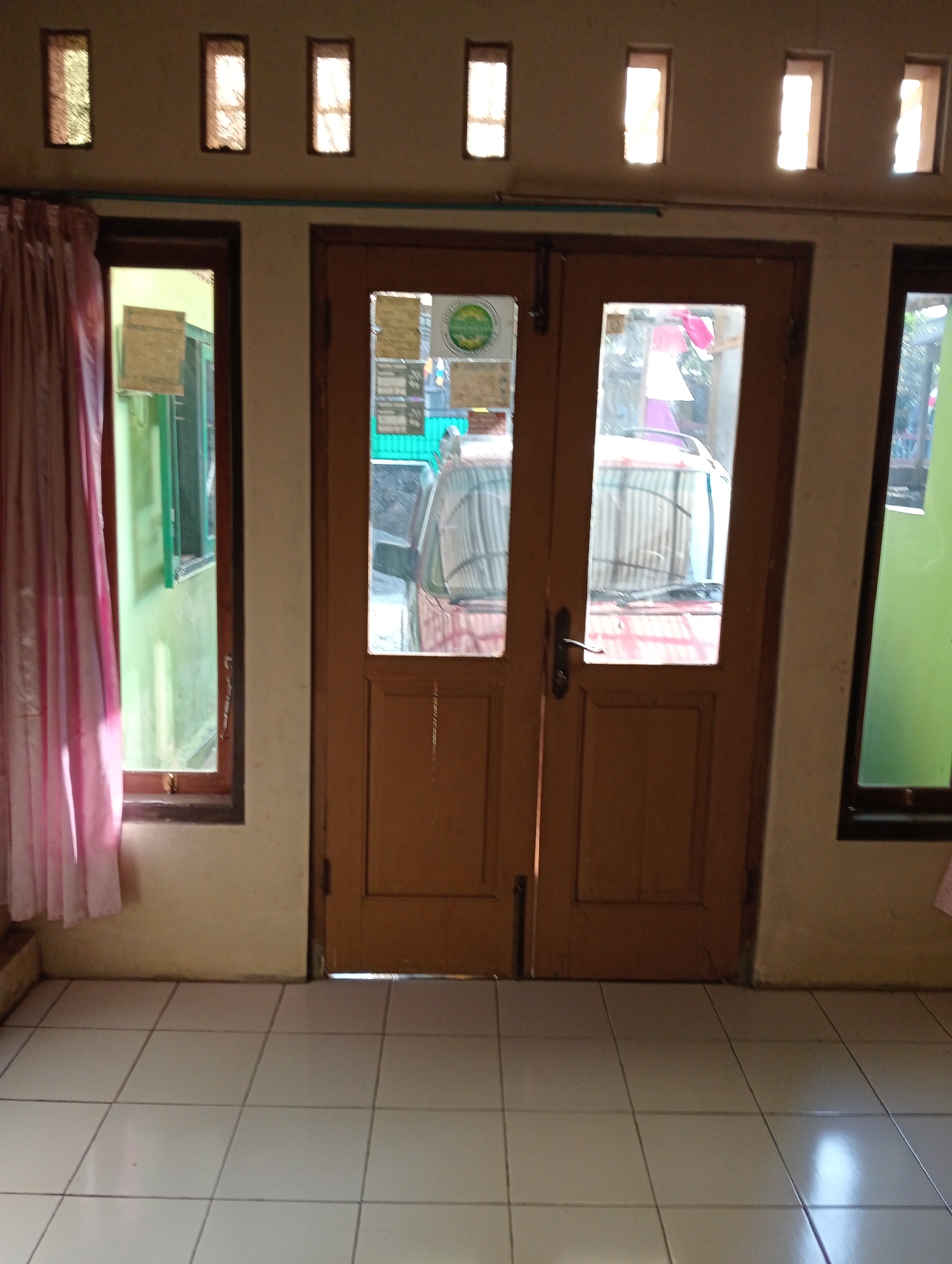 The image depicts a frontal view of a double wooden door, painted a medium brown, centrally located within a room. Each door has two rectangular glass panes, revealing glimpses of a red car with a corrugated metal roof parked outside. The door handles are simple metal levers. To the left of the doors, a window with a dark wooden frame is partially covered by a light pink curtain. To the right of the doors, another window with a dark wooden frame is visible. Above the doors and windows, there are four rectangular openings in the wall, serving as ventilation. The floor is tiled with large, square white tiles. The walls are painted a light beige. The scene appears to be indoors, facing outwards, likely in a residential or small commercial setting in Krajan Kidul, Indonesia, given the context.