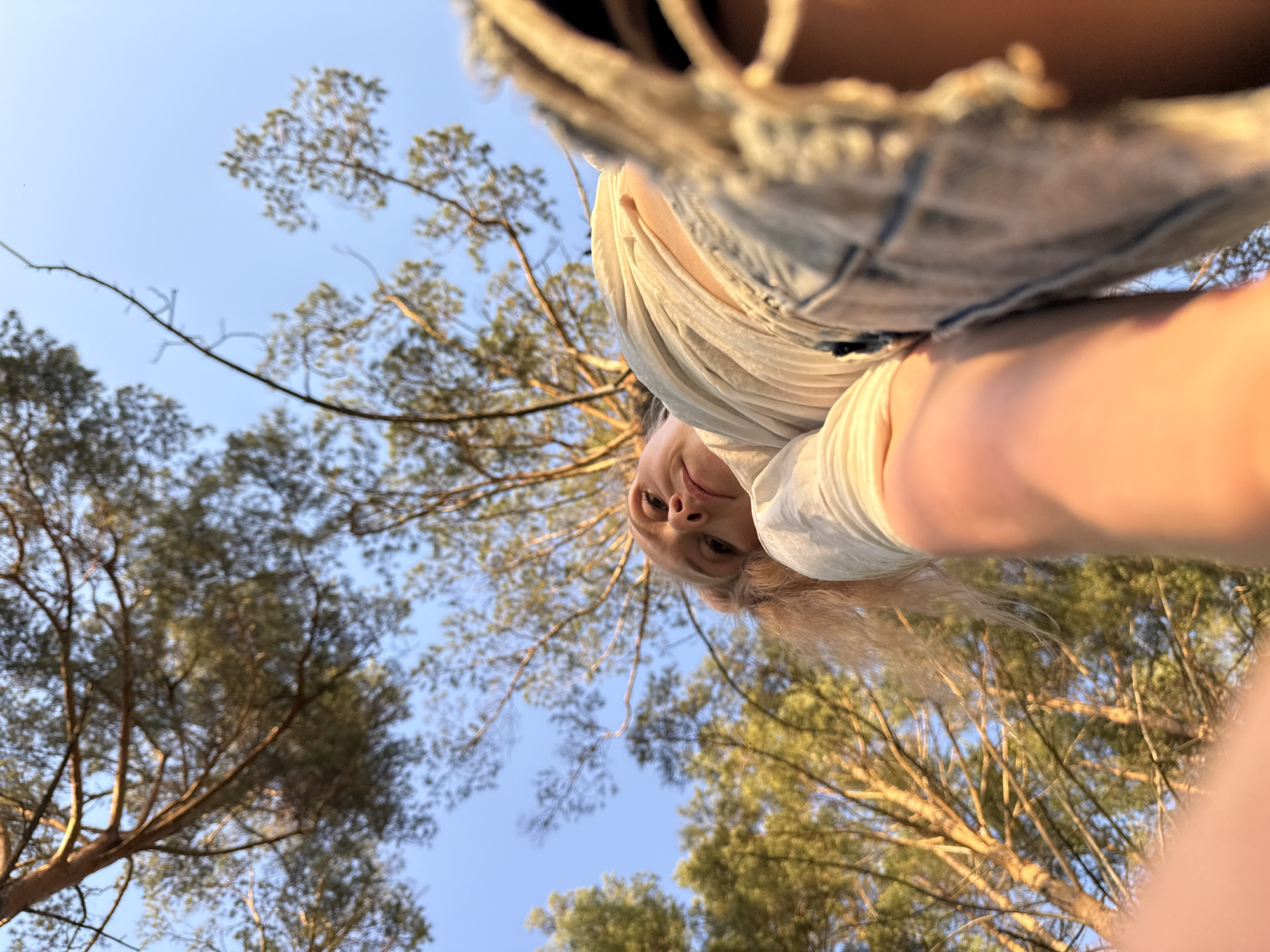 The image, captured from a low angle, depicts a woman amidst a backdrop of pine trees and a clear blue sky. She wears a light-colored shirt and denim shorts. The location is potentially a forested area within or near Прикамский, Russia, indicated by the presence of pine trees.