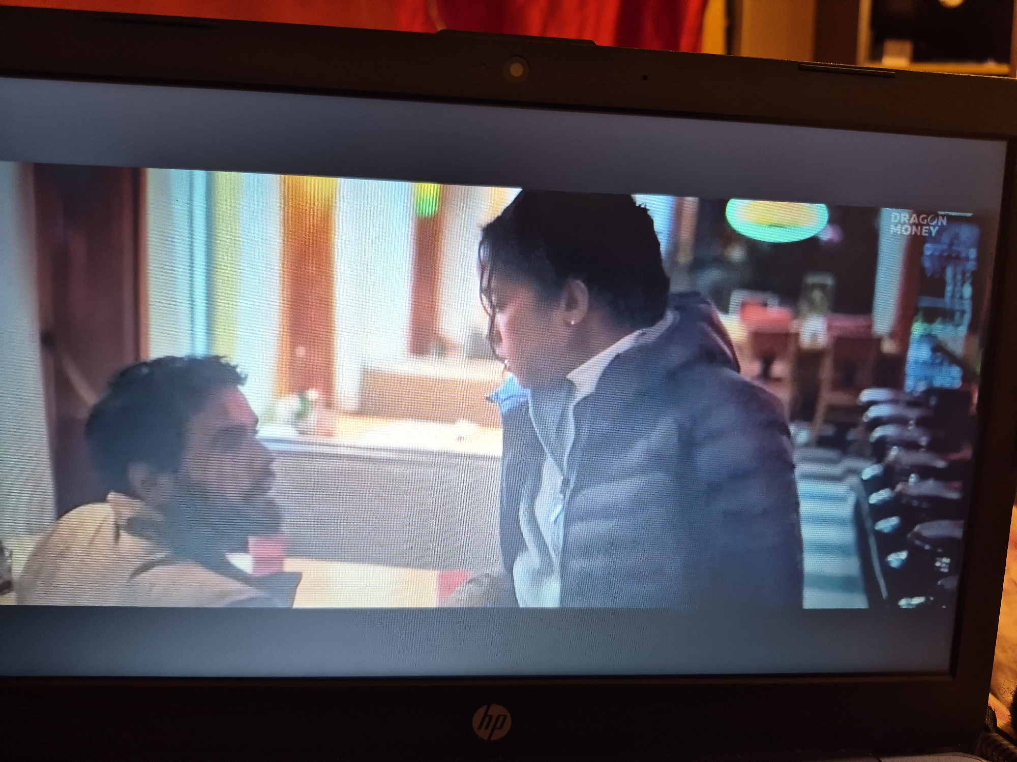 The media file shows a scene from a laptop screen, displaying a video. Two individuals, a man and a woman, are the subjects. The man, on the left, has dark hair and a beard and is wearing a light-colored shirt. He is looking towards the woman, and his expression appears serious. The woman, on the right, has dark hair and is wearing a puffer jacket over a white shirt. She is also looking towards the man, and her expression suggests an intense conversation. They appear to be in an indoor setting, possibly a diner or restaurant, with booth seating visible. The background features vertical colored panels, suggesting a stylized interior. A sign with "DRAGON MONEY" is visible in the upper right corner of the video, indicating a potential business or brand. The lighting suggests it could be evening or an indoor environment with artificial lighting. The overall mood of the scene seems to be one of a serious or dramatic interaction between the two people.
