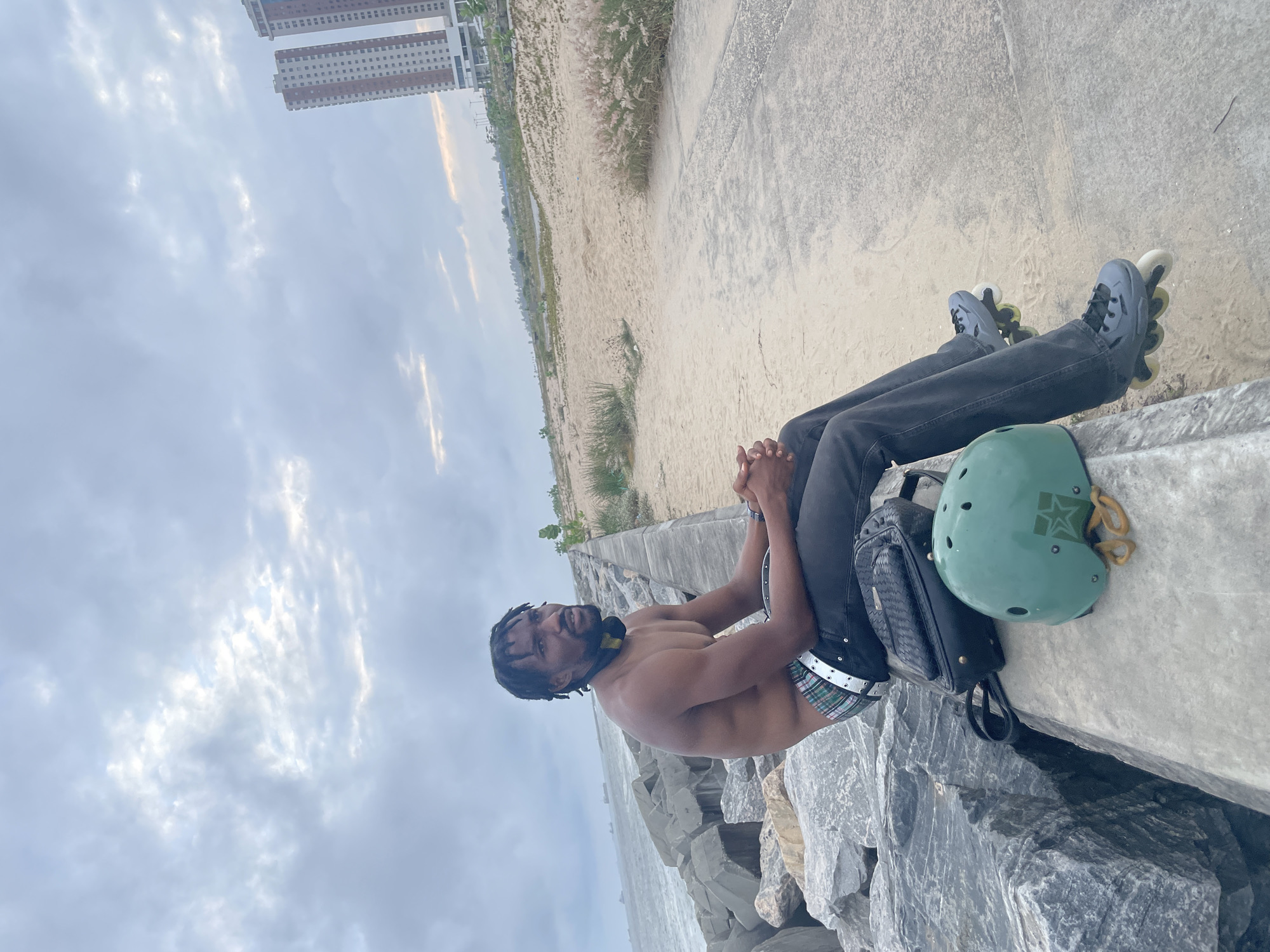 A shirtless Black man with dreadlocks, wearing dark jeans and rollerblades, sits on a concrete wall by the ocean in Inupa Kekere, Nigeria. He has a black bag and a green helmet beside him. The background shows a beach, high-rise buildings, and ships in the distance. The sky is overcast with scattered clouds.