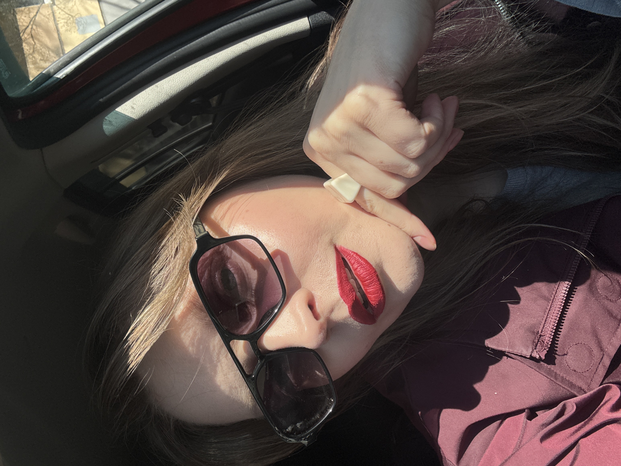 The image presents a close-up, high-angle shot of a person lying down inside a car. The primary subject is an individual with long, light brown hair, bold red lipstick, and black-framed sunglasses resting on their nose. One hand, adorned with a distinctive white, geometric ring on the index finger, is raised towards their face, partially obscuring it. The person is wearing a burgundy or maroon garment, possibly a jacket, which features a visible zipper.
Strong, direct sunlight illuminates parts of the face, hair, and hand, creating stark highlights and pronounced shadows. This indicates a sunny day, likely midday or afternoon. The car's interior is visible, including the door frame, a window showing blurry outdoor scenery, and the light-colored roof lining. The overall composition suggests a stylized pose within a casual setting. There is no visible text in the image. This photograph was taken in Izhevsk, Russia.