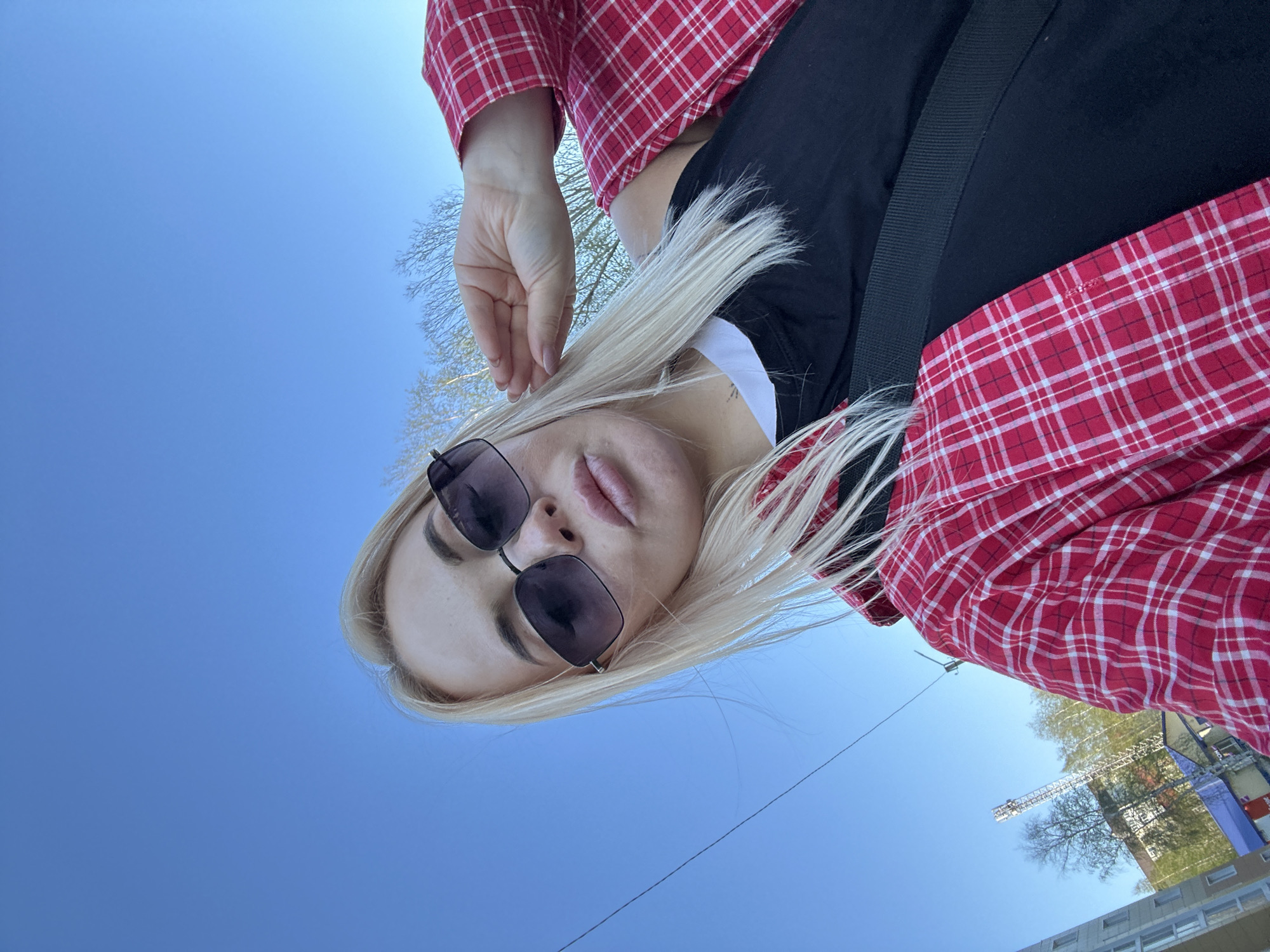 A woman with blonde hair and sunglasses is viewed from a low angle against a bright blue sky. She is wearing a red and white plaid shirt over a black top, with a white collar visible. A black strap crosses her body. Her hand is raised near her head. In the background, a building with blue reflective glass, trees, and a white construction crane are visible. The location is likely Megion, Russia, as indicated by the crane and building style. The image is taken outdoors in daylight.