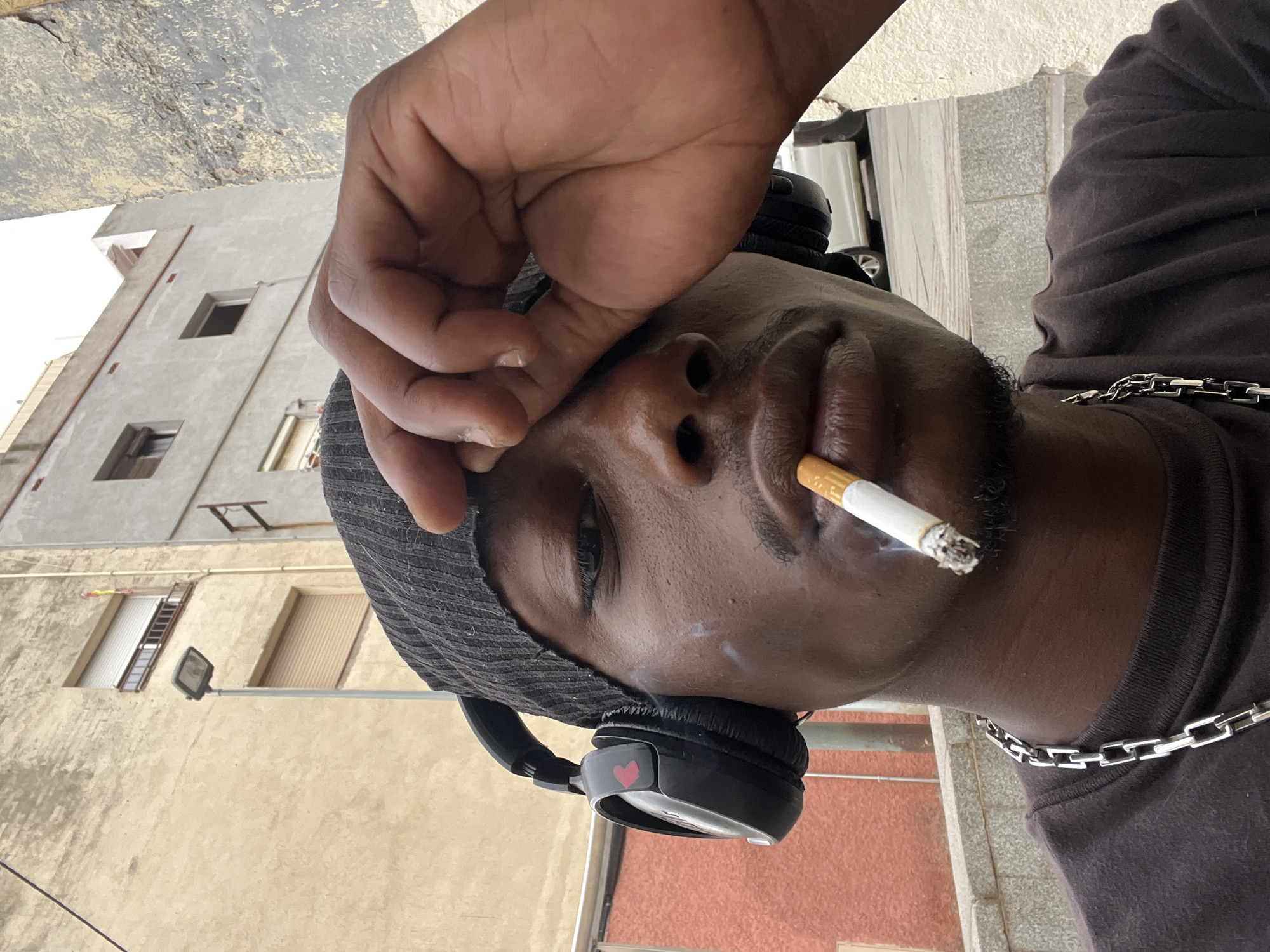 A man with dark skin is holding a lit cigarette in his mouth. He is wearing a dark-colored beanie and headphones with a red heart on one side. A silver chain is visible around his neck. His hand is raised towards his forehead. The background shows a building with windows and a street lamp, suggesting an urban environment. Based on architectural style and coloring, it is possibly in the city of Barcelona, Spain.
