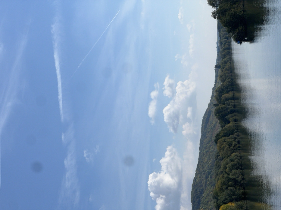 The image displays a wide landscape scene of the Seine River near Vernon, France, observed under a bright blue sky. The sky contains scattered white cumulus clouds, wispy cirrus clouds, and two distinct aircraft contrails: one long, thin, and diagonal across the upper right, and another broader, fading one in the upper left. Several dark, circular artifacts, indicative of lens flares or sensor dust, are visible against the sky.
The background consists of rolling hills covered in dense, dark green deciduous forest, with some trees showing hints of yellowing foliage, suggesting an autumn season. The wide, calm river occupies the mid-ground and foreground, reflecting the blue sky and the trees on its banks. Both river banks are extensively lined with mature trees. On the right side of the river, positioned near the bank amidst the trees, a small, dark-colored object, possibly a boat or a fixed structure, is visible with a blue covering or tarp on its upper surface. Small, dark specks, consistent with distant birds, are sparsely distributed in the sky. There are no discernible human figures, actions, or interactions within the frame.