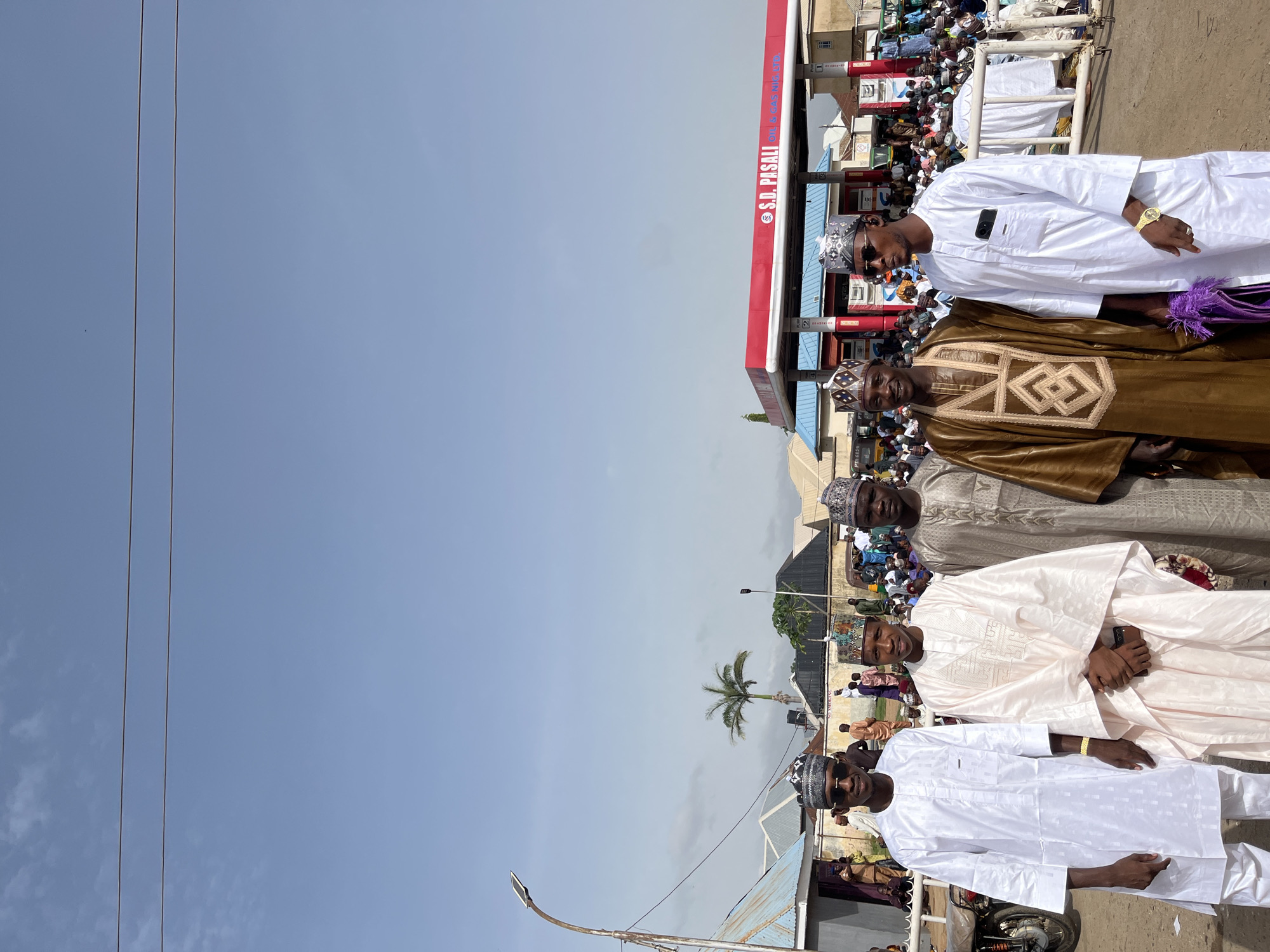 The image captures a group of five men standing close together, possibly posing for a photo. They are all dressed in traditional clothing with intricately designed hats. The first man on the right wears a white robe and sunglasses, while the second is dressed in a brown robe with an elaborate design on the front. The third man wears a beige robe, and the fourth is in a light pink robe. The fifth man on the left is again wearing a white robe and sunglasses.
Behind the men, there is a large crowd of people. In the background, a gas station with red and blue signage is visible. Further back, a palm tree can be seen amidst buildings and power lines. The sky above is clear and blue.