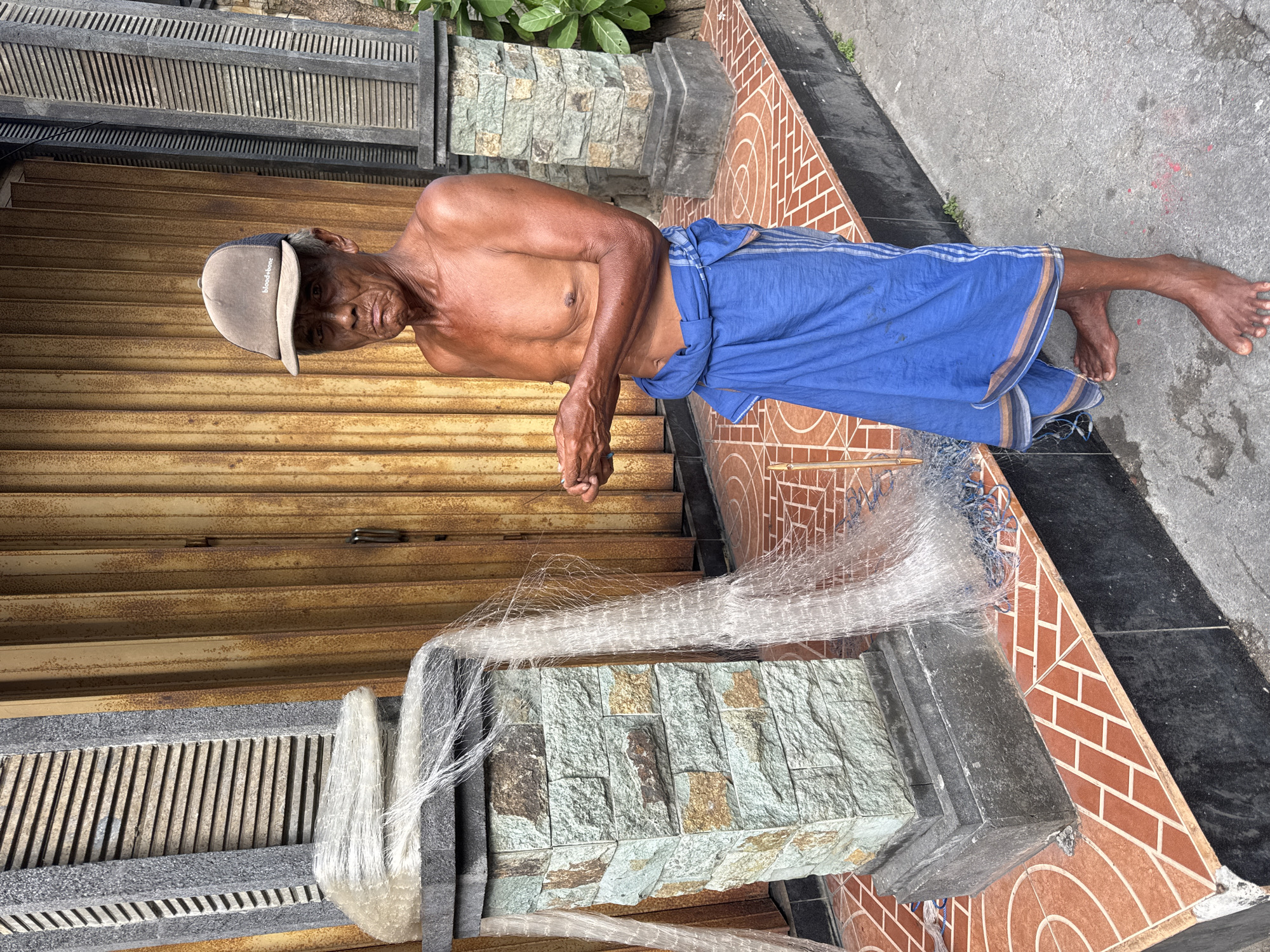 An eye-level medium shot of an elderly man standing near a building with a fishing net, taken in Kuta, Indonesia.
The man is dark-skinned and shirtless, wearing a blue sarong wrapped around his waist and a light-colored baseball cap. He is holding a fishing net in his hands.
The building has a rusted, corrugated metal door and decorative stone accents. The foreground features a stone pillar, draped with the same fishing net, standing on a patterned tile surface. A concrete sidewalk extends towards the top-right corner of the frame. A narrow strip of green vegetation is visible along the top edge of the frame.
The lighting is slightly overcast. The image seems to be taken during the day. The camera angle is vertical.