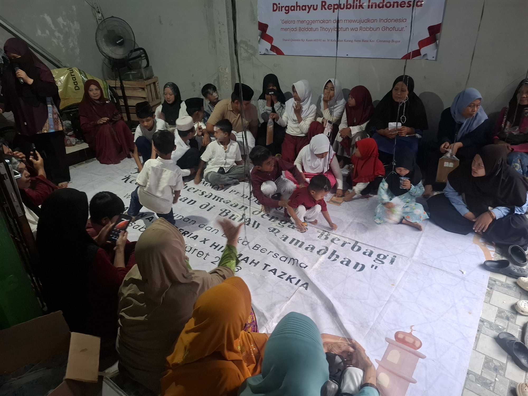 In a dimly lit indoor space, likely a community hall or room in Babakan Madang, Indonesia, a gathering of approximately 20 people, predominantly women and children, are seated on the floor around a large white banner. The banner features text in Indonesian and a graphic of a mosque tower, suggesting a religious or community event. A ceiling fan is visible in the background. Many individuals are wearing headscarves, common attire for women in Indonesia. Some children are interacting with each other, while adults appear to be observing or listening. One woman on the left is holding a microphone, possibly addressing the group. The overall atmosphere seems to be one of communal gathering and cultural engagement.