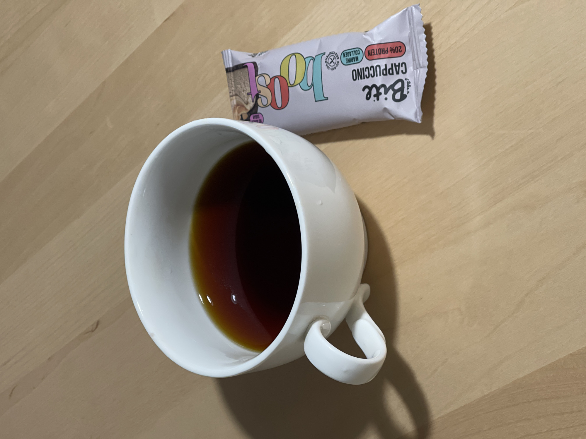 A top-down shot shows a white ceramic mug containing a dark liquid, possibly coffee or tea, next to a protein bar on a wooden surface. The protein bar is labeled "Bite Cappuccino 20% Protein Marine Collagen." No people are visible. The image does not suggest a specific location in Izhevsk, Russia, or any other location.