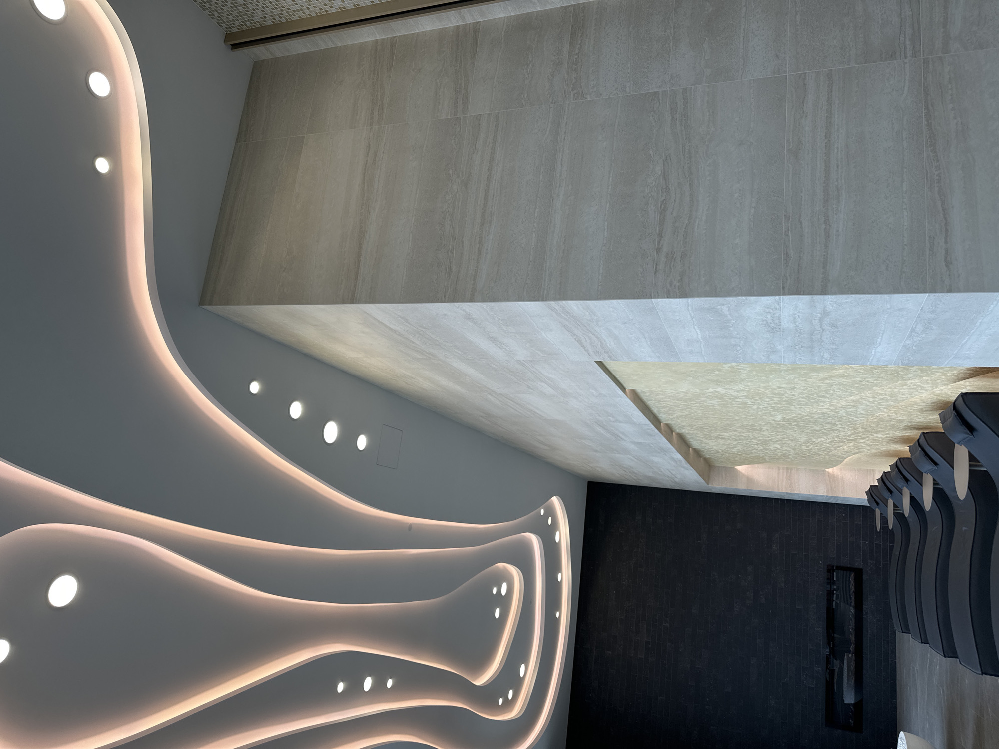 The image captures an interior space with a modern and stylized design. The ceiling is a prominent feature, characterized by wavy, illuminated structures with embedded round lights, creating a soft, ambient glow. The walls are covered with light-colored, horizontally laid stone tiles with a subtle, linear pattern, giving them a textured appearance. The floor has a darker, vertically aligned brick-like covering. A row of sculpted, dark chairs stands against the wall. The photograph's location is not possible to determine based on the image alone.
