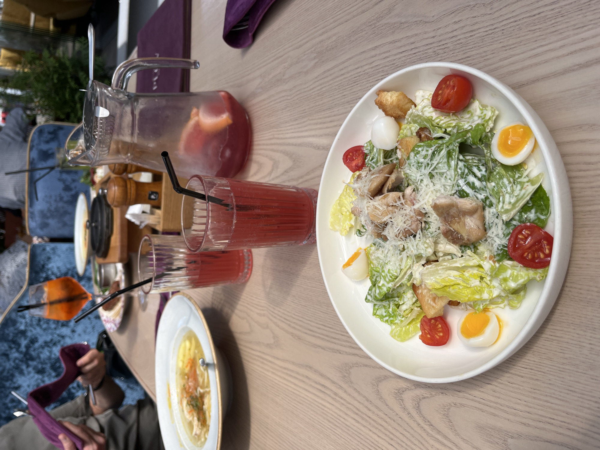 Here is a detailed description of the image:
**Overall Impression:** The image depicts a table setting at a restaurant, showcasing a meal in progress. The scene seems to be taken from an elevated perspective, possibly by someone sitting at the table.
**Objects and Food:**
* A plate of what appears to be a Caesar salad with chicken, croutons, parmesan cheese, eggs and tomatoes.
* Two glasses of red liquid with straws
* A pitcher containing a red liquid with visible fruits
* A bowl of what appears to be chicken soup
**Location Details:**
* It is difficult to pinpoint the exact location in Yekaterinburg, Russia, from this image alone.
**People:**
* Partial view of a person holding a napkin, wearing a watch, holding silverware, and holding a purple cloth.
**Environment:**
* The table is a light-colored wooden table with a neutral, possibly lacquered, finish.
* The lighting appears to be a mix of natural and artificial, creating a well-lit setting.
**Actions/Interactions:**
* The person is wiping their hands or mouth with the napkin after eating.
* The presence of the food and beverages indicates a meal in progress.