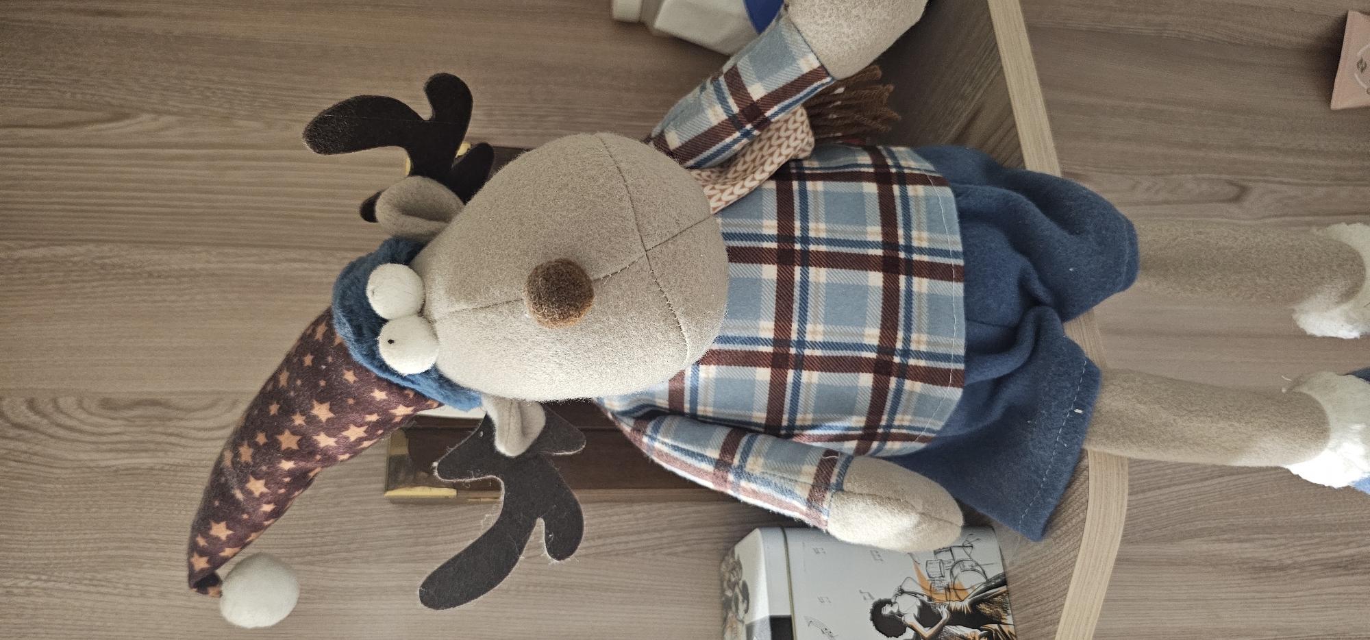 A stuffed reindeer toy is seated on a wooden shelf in an indoor setting. The toy is wearing a festive, pointed hat with stars, a blue knitted hat, a plaid shirt with blue, brown, and white checkered patterns, blue shorts, and white fuzzy trim on its legs. It has large, googly eyes and antlers. To the left of the toy is a decorative tin box featuring an illustration of a woman playing a musical instrument. The background consists of a wooden shelf with vertical grain. The scene is well-lit, likely from natural light sources. The exact location within Vinnytsia, Ukraine, cannot be determined from the image.
