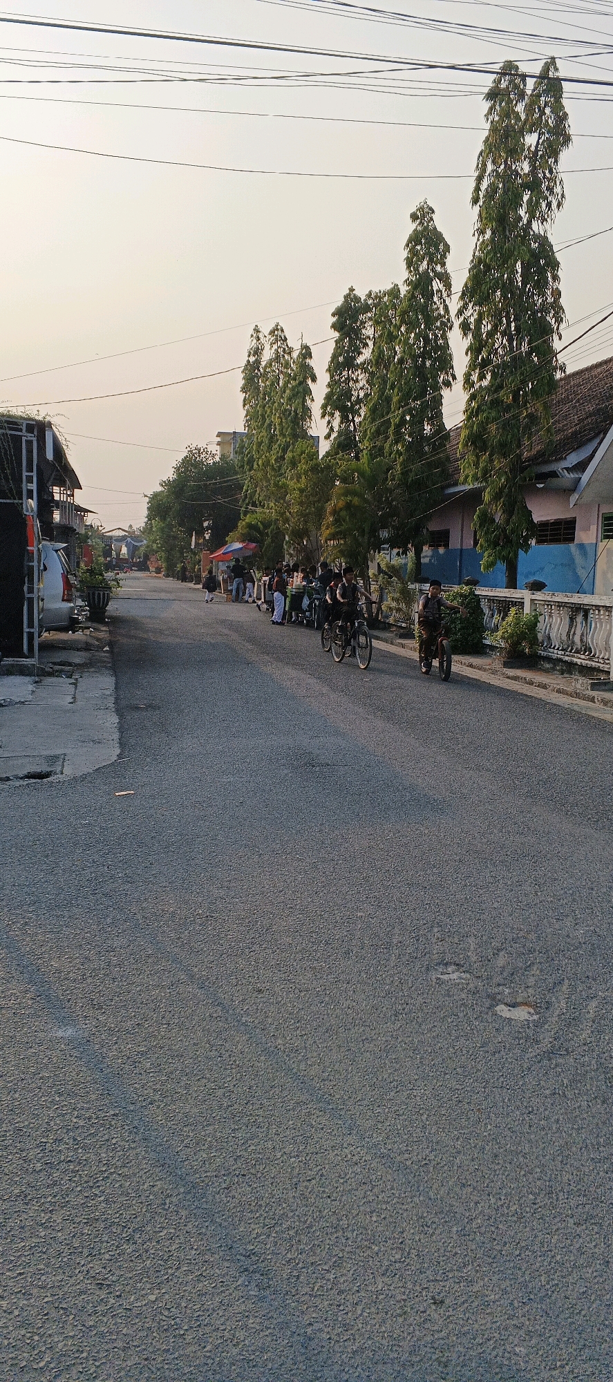 The image depicts a residential street scene in Latukan, Indonesia. The street is paved with asphalt and is lined with houses and tall trees. In the foreground, a young person is riding a bicycle, and another person is seen on a motorcycle in the distance. There are several other individuals gathered on the side of the road, some appearing to be standing or sitting. Several parked vehicles, including a silver car and a motorcycle, are visible on the left side of the street. Overhead, a complex network of electrical wires stretches across the sky. The lighting suggests it is late afternoon or early evening. The environment appears to be a typical Indonesian urban or suburban setting.