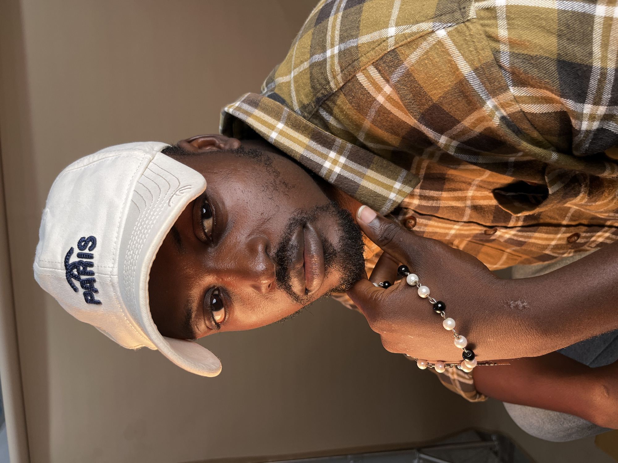 The image shows a close-up portrait of a Black man wearing a white baseball cap with "PARIS" embroidered on it in navy blue. He has a beard and is looking directly at the camera with his left hand resting on his chin, holding a bracelet made of black and white beads. He is wearing a brown and yellow plaid shirt. The background is a plain, warm-toned wall with some lighting effects. The image appears to be taken indoors, possibly in a studio or residential setting. There is no visible indication of the specific location being Gura Top, Nigeria.