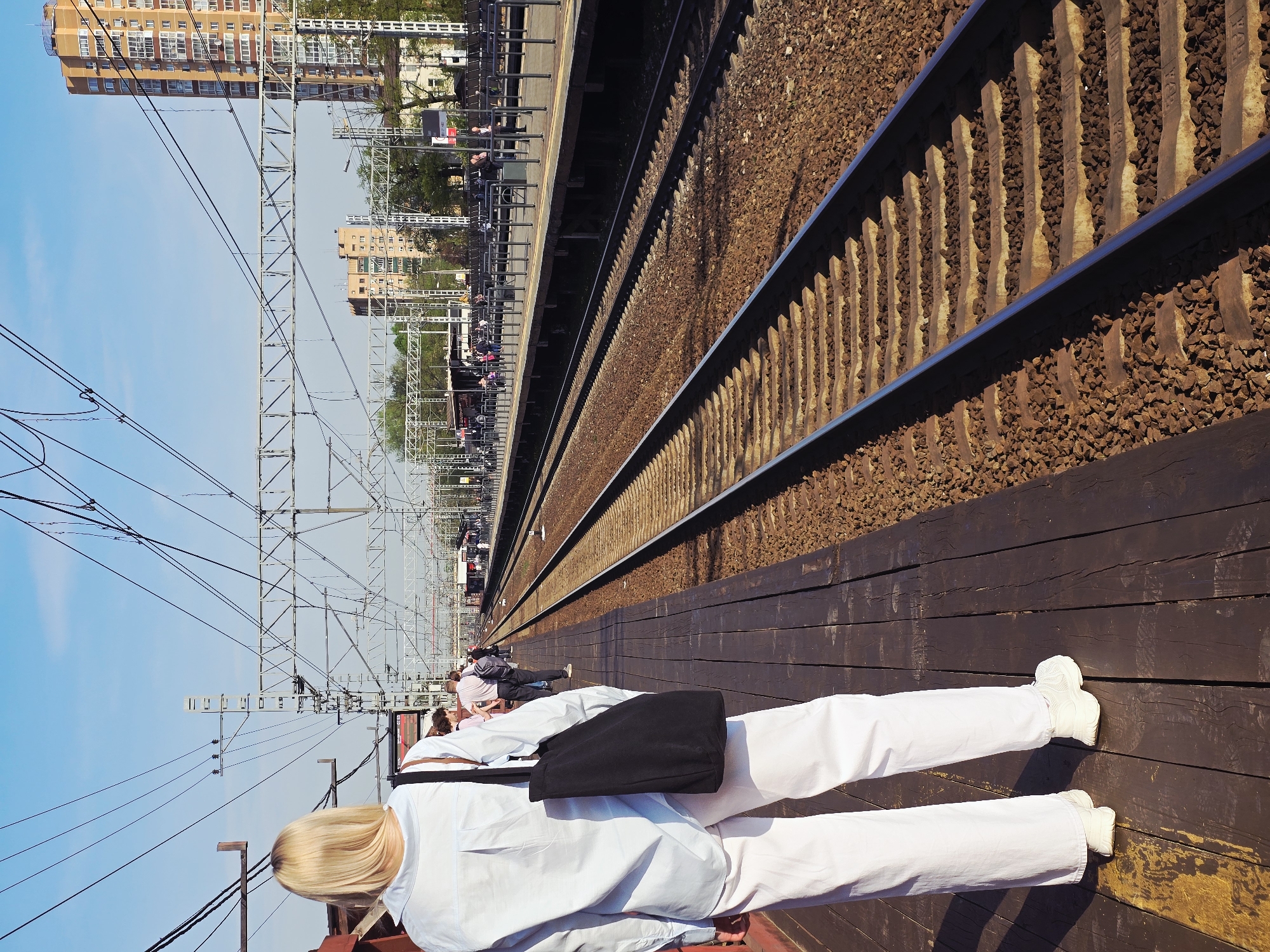 The image depicts a view from a train platform in Khimki, Russia. The scene shows a railway track with ballast, wooden sleepers, and metal rails, extending into the distance. A wooden platform borders the track. A person with blonde hair and wearing white clothing and white sneakers stands on the platform with a black bag slung over their shoulder. Other people are visible in the distance on the platform. Overhead power lines and supporting structures stretch across the sky. Buildings and trees are visible in the background, suggesting an urban environment. The sky is blue.