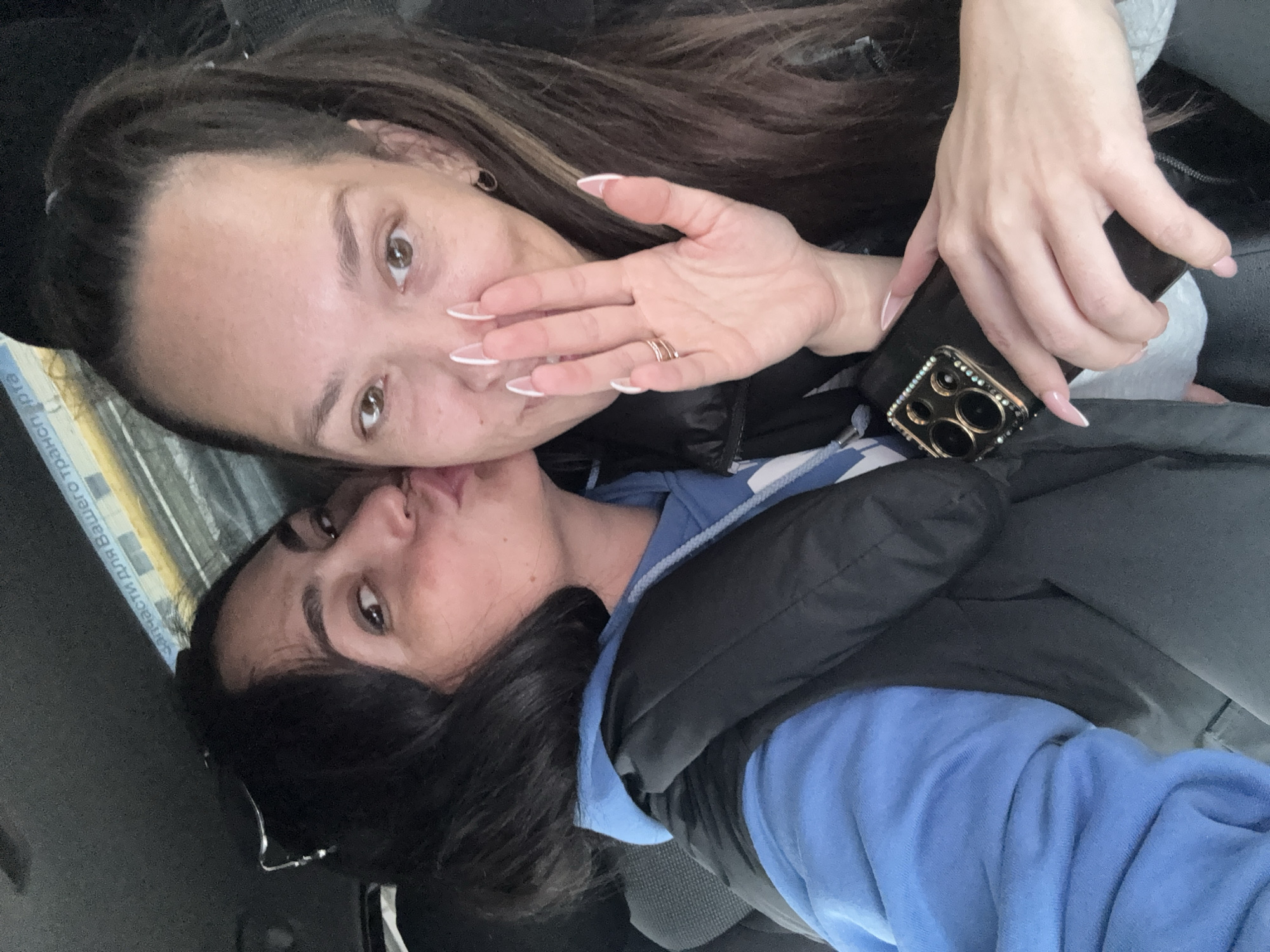 This rotated selfie captures two women in what appears to be an indoor or vehicle setting, with the provided context indicating Chaikovsky, Russia. The woman positioned at the top of the frame, likely holding the camera, has dark hair, dark brown eyes, and is looking directly forward. Her right hand, adorned with long, white-tipped nails and a ring on her ring finger, is partially covering her mouth. She holds a smartphone encased in a decorative cover featuring rhinestones around the camera lenses.
Below her, the second woman, also with dark hair and dark brown eyes, is making a playful kissing face with pursed lips. She is wearing a bright blue hooded sweatshirt layered under a dark, possibly black, puffer vest or jacket. The overall expression conveyed by both individuals suggests a friendly or affectionate interaction.
The background is mostly dark and indistinct, consistent with a car interior or similar confined space. On a blue and white patterned object in the upper left background, partially visible Russian text reads "Запчасти для Вашего транспорта," which translates to "Spare parts for your transport," reinforcing the likelihood of a vehicle setting.