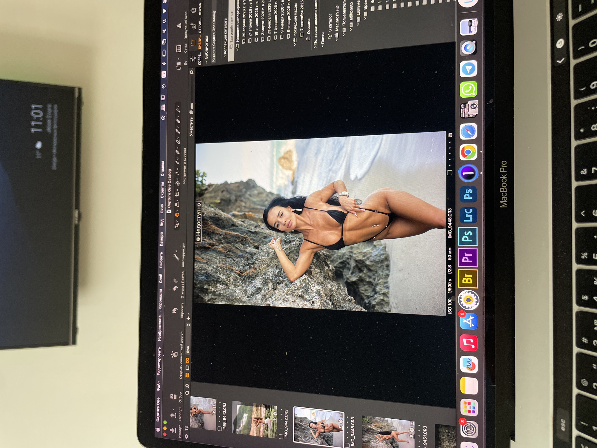 A laptop screen displays a photo editing program (Capture One) with a woman in a black bikini posing on rocks near a beach. The laptop is a MacBook Pro, with the keyboard visible. The screen also shows open applications (Photoshop, Premiere Pro, etc.) and dock icons. The date shown is April 29th, 2026. A clock in the upper left shows the time as 11:01. The environment is an office setting, with a TV screen partially visible on the left. There is no indication of this image being in Kazan, Russia.