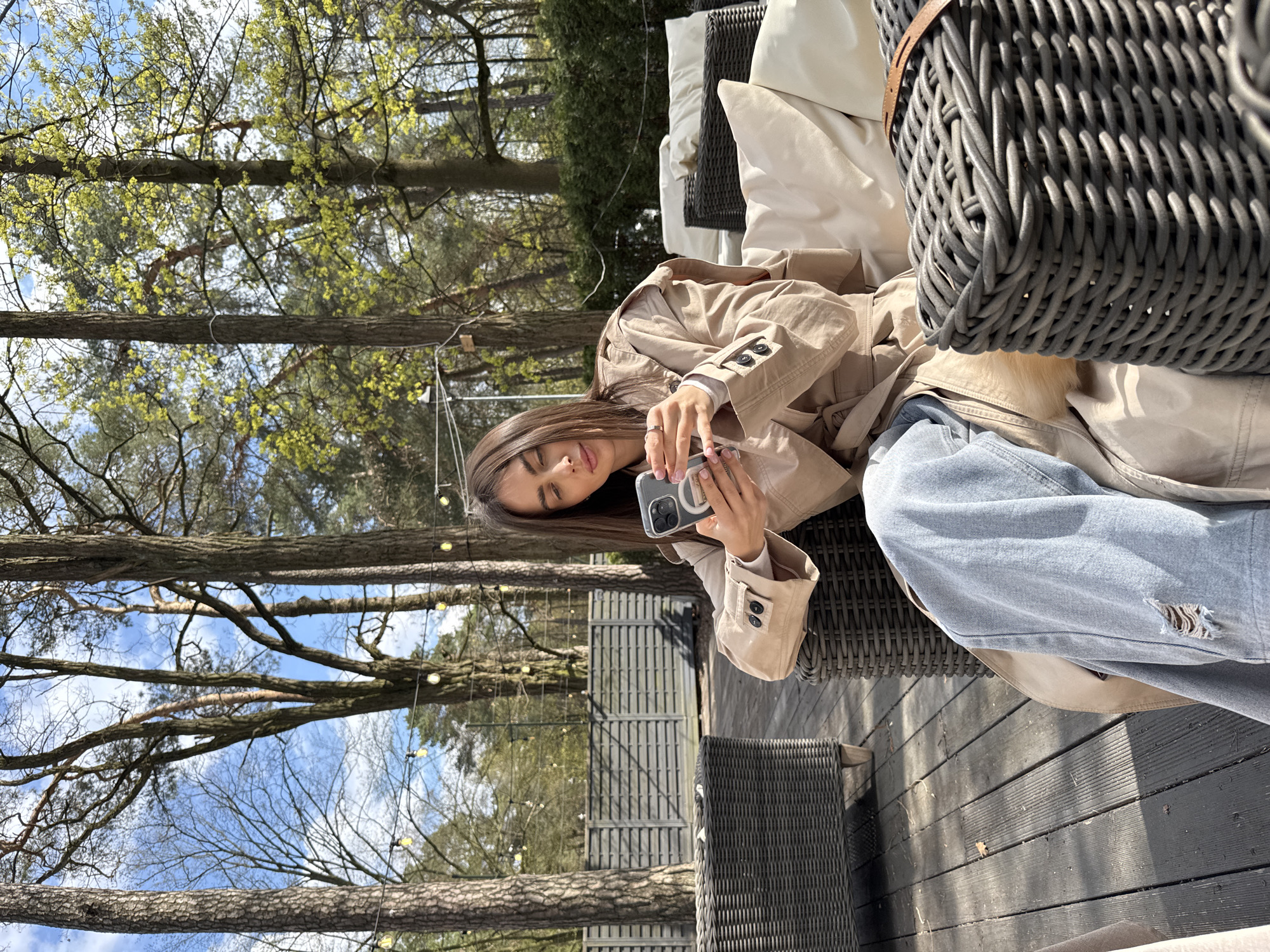 A woman with long brown hair is sitting on a wicker sofa in what appears to be an outdoor setting. She is wearing a tan trench coat over a white top and ripped light blue jeans. She is holding a smartphone in her hands. A large grey wicker bag is positioned to her right, partially obscuring the sofa. The background shows trees with sparse leaves, hinting at early spring. Strings of small lights are strung across the tree branches. The wooden deck on which the sofa sits indicates an outdoor patio or garden setting. Based on the visual information, the specific location within Konstancin-Jeziorna, Poland, cannot be definitively determined. However, the scene depicts a relaxing outdoor setting possibly in a garden or patio space, suggesting a residential or leisure area.