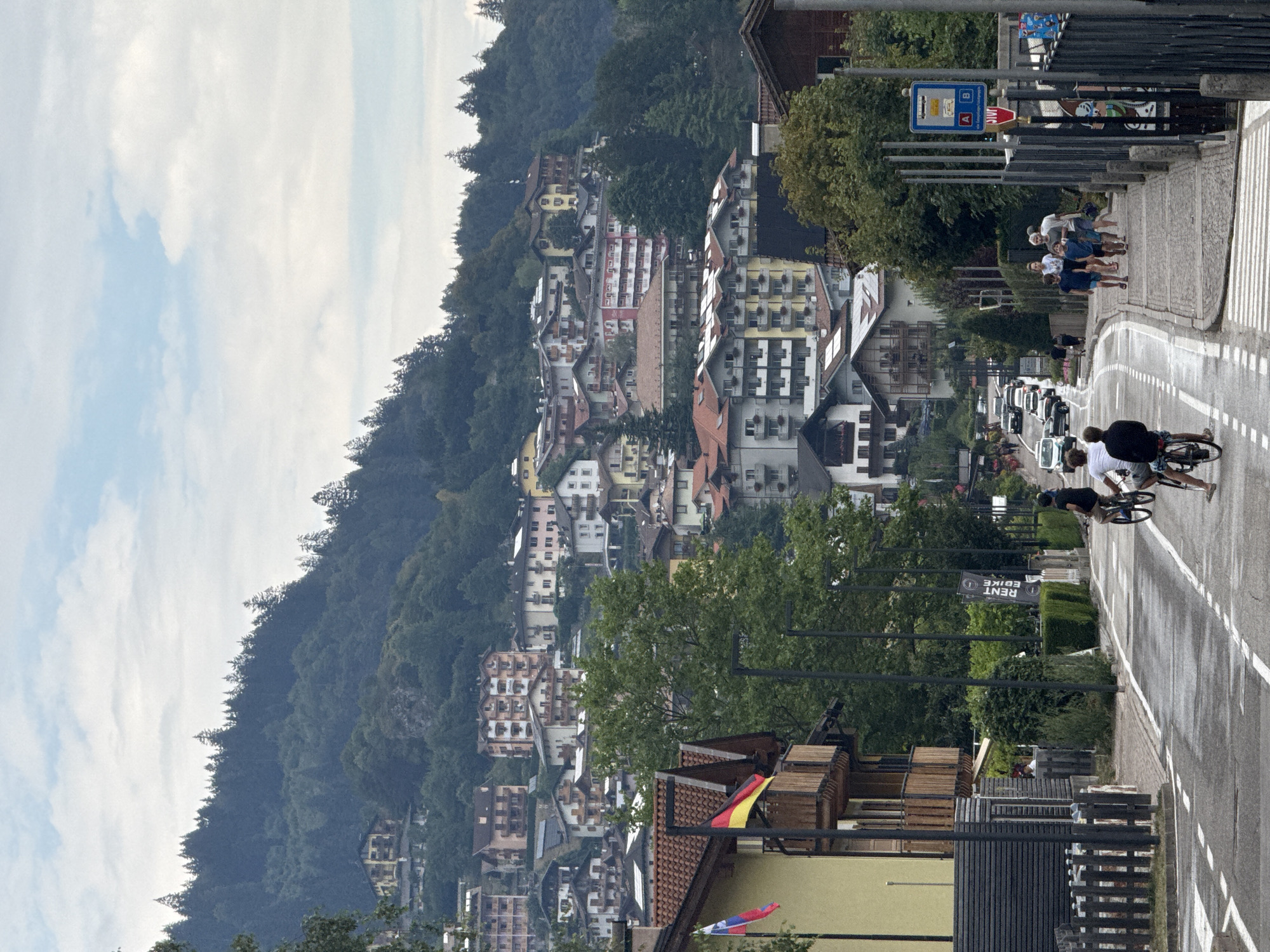 The image depicts a street scene in the Italian city of Molveno, with a steep hill covered in dense green trees in the background. Numerous multi-story buildings with red-tiled roofs and balconies are built into the hillside, characteristic of mountain towns. In the foreground, a paved road curves upwards, lined with sidewalks and streetlights. Several people are visible on the sidewalk and road, some cycling and others walking. Two flags, one Slovenian and one German, fly from a balcony on a yellow building on the left. A bus stop sign is also visible on the right. The overall atmosphere suggests a tranquil day in a picturesque European mountain town.