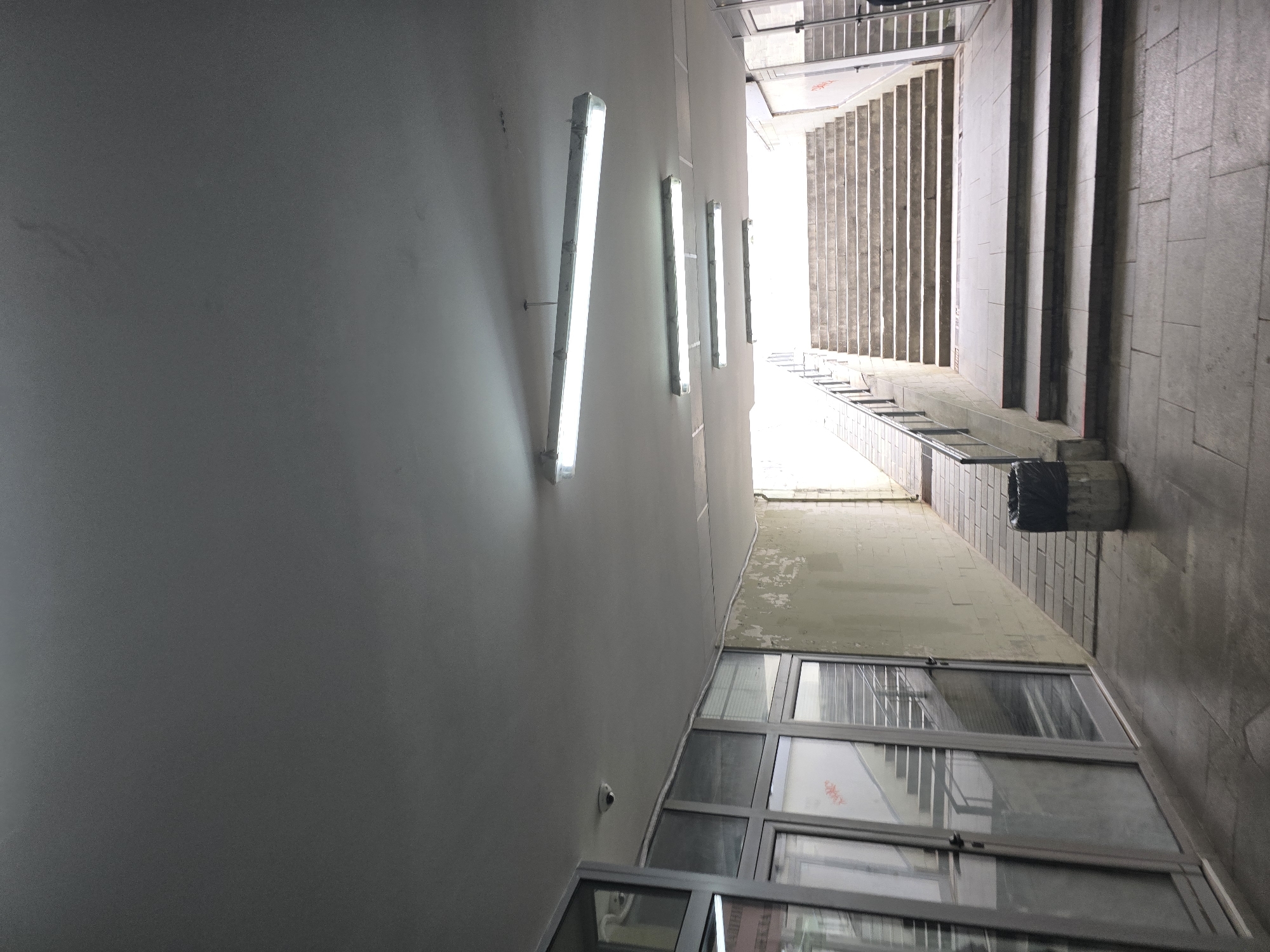 The image shows an upward-angled perspective of a narrow corridor or passageway, likely in a building in the city of Unknown, Unknown. The left wall is a smooth, light gray, with four fluorescent tube lights mounted along its length. The right side of the passageway features a series of windows near the bottom and a set of concrete stairs leading upwards towards a bright opening. A trash receptacle sits near the base of the stairs. The angle of the shot provides a sense of depth and emphasizes the verticality of the space.