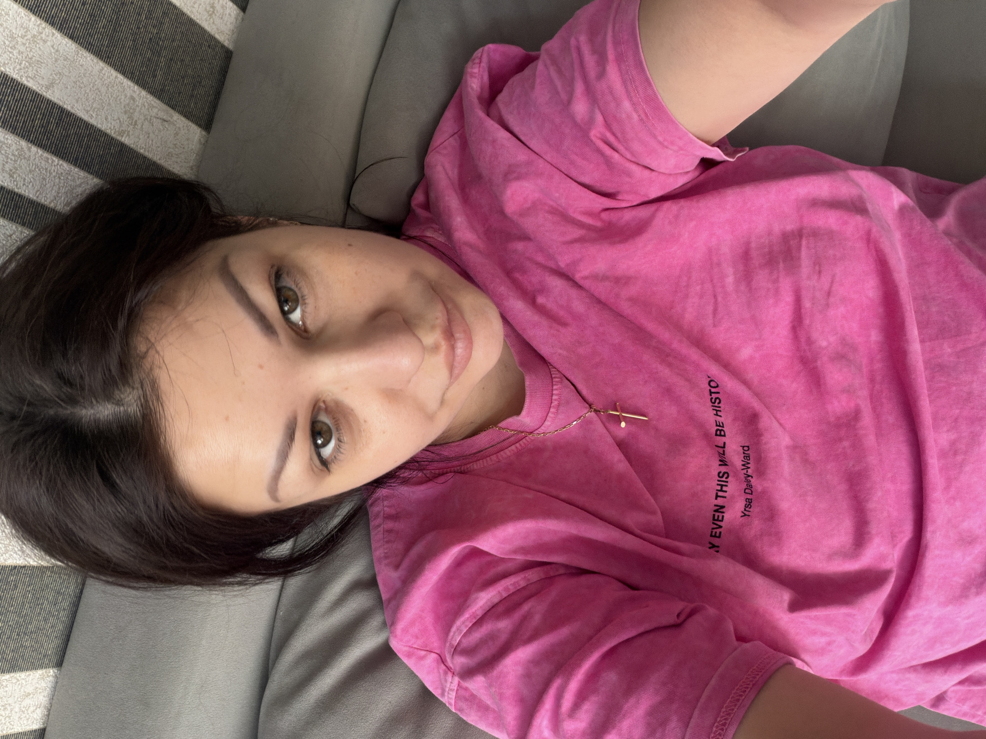 A young woman with dark hair and brown eyes lies on a gray sofa, looking upwards at the camera. She is wearing a bright pink t-shirt with a gold cross necklace. The t-shirt has text printed on it, which reads "Y EVEN THIS WILL BE HISTO" and "Yrsa Daley-Ward". The background consists of a striped pattern on what appears to be a headboard or wall. There are no indications of the city of Sochi, Russia in the image.
