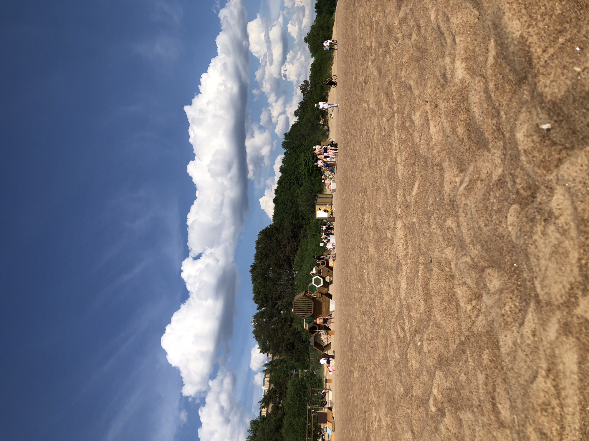 This wide-angle photograph depicts a sandy beach under a bright blue sky with numerous scattered white cumulus clouds in Sestroretsk, Russia. The foreground consists of light brown sand exhibiting discernible ripple patterns. In the middle ground, multiple individuals are present. A prominent wooden play structure, composed of interconnected hexagonal modules with some green and white accents, stands centrally, with people interacting around and on it. A shirtless adult male in dark shorts is positioned directly in front of this structure. To the left, smaller wooden structures, including ramps, are visible near other individuals. To the right, a cluster of people gathers near a small, light-colored rectangular building. Individuals are observed walking, standing, sitting, and engaging with the play installations. At least two people are depicted riding bicycles across the sand in the mid-right. A dense treeline, comprising both coniferous and deciduous trees, forms the background, delineating the rear boundary of the beach. The scene is illuminated by strong sunlight, indicating clear weather.