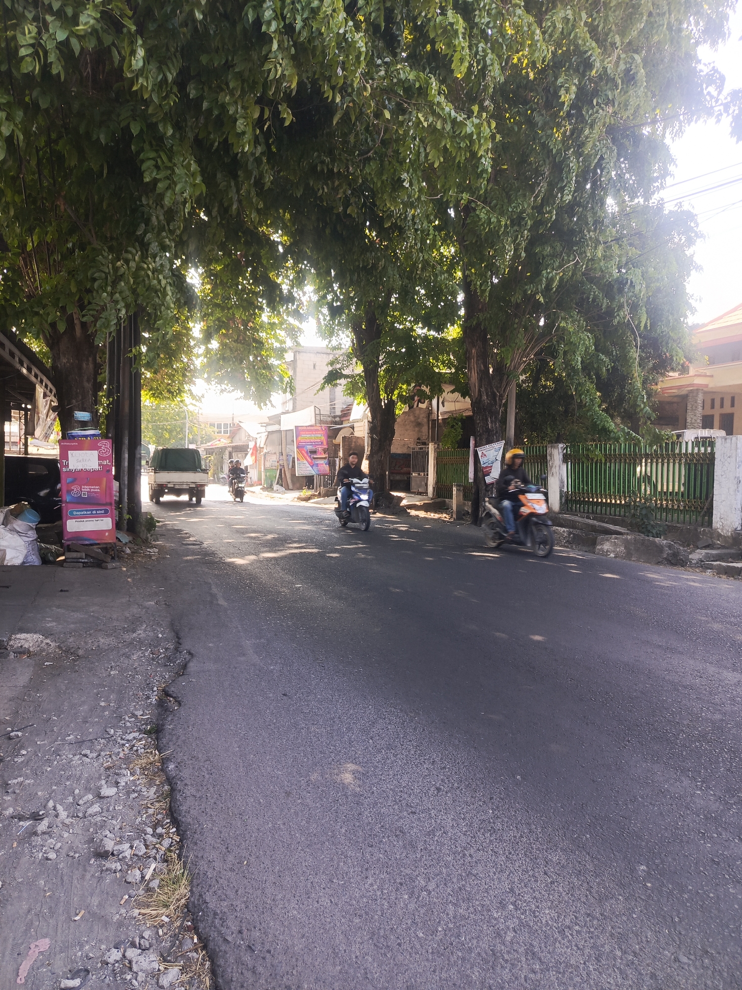 The image depicts a street scene in Sidowungu, Indonesia, under a clear sky with sunlight filtering through lush green trees lining the road. The road itself is paved with asphalt and shows signs of wear, with a rougher, gravelly shoulder on the left side.
Several people are present, all riding motorcycles. In the foreground on the right, a person wearing a yellow helmet and an orange and black jacket rides an orange and black motorcycle. Further up the road, another person in a dark jacket and jeans, also wearing a helmet, rides a white motorcycle. In the distance, a person in a dark shirt and blue pants on a dark motorcycle is visible. To the left, a white truck with a green tarp covering its cargo is parked or moving slowly.
Along the left side of the road, a building with a dark roof is partially visible, and a tall, slender sign with colorful graphics and text is displayed. Bags of what appear to be trash or materials are piled near the sign. The right side of the street features a green metal fence with white concrete posts, bordering a building with a light-colored facade and a tiled roof. Overhead, electrical wires are strung across the scene. The overall impression is a typical day on a residential or commercial street in a tropical climate.