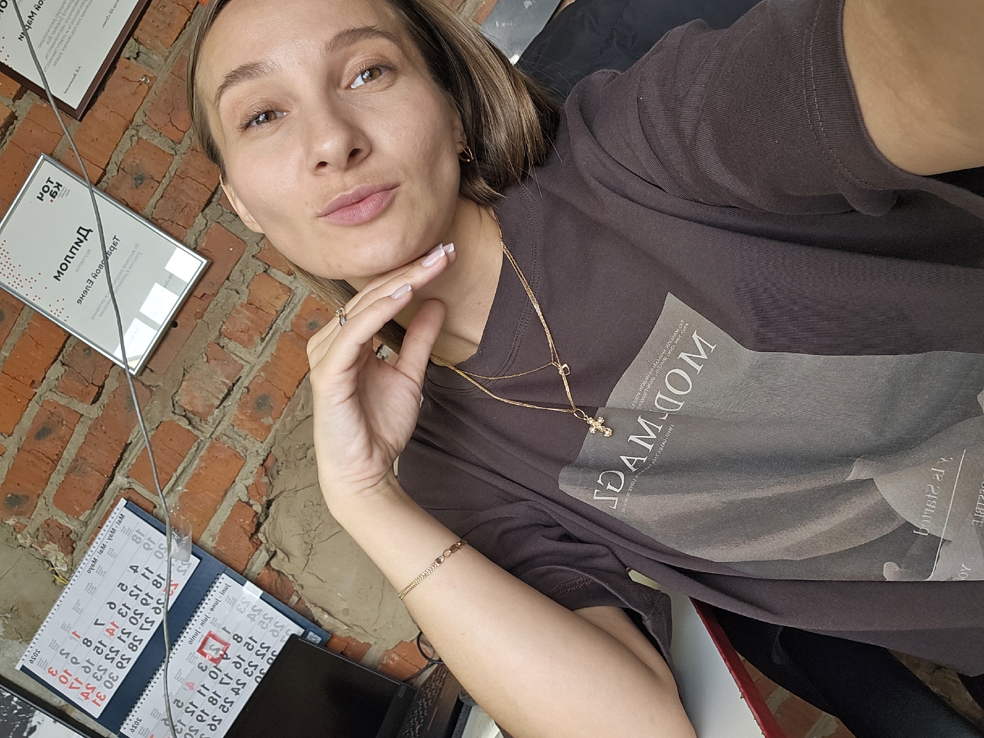 A young woman poses for a selfie indoors in Chaikovsky, Russia. She has light brown hair, warm brown eyes, and is looking directly at the camera with a relaxed expression, her lips slightly pursed. Her right hand is gently placed under her chin, revealing light-colored nails with white tips, a ring on her finger, and a delicate gold bracelet on her wrist. She wears a dark brown, oversized t-shirt with a light-colored graphic print featuring text that includes "DAM.COM" and possibly "MODDAM". Multiple delicate gold chain necklaces, one with a small cross pendant, and small gold hoop earrings complete her accessories.
The background features a rustic red brick wall, lending an industrial or cozy feel to the setting. Two framed documents or certificates are displayed on the wall, one clearly visible with Russian text including "МОЛЛПД", "Диплом" (Diploma), and a name "Елене Эдуардовне" (Elena Eduardovna), indicating an award or achievement "За участие в Всероссийской" (For participation in the All-Russian...). Below these, two small, spiral-bound desktop calendars in Russian show months like "Октябрь" (October), "Ноябрь" (November), and "Декабрь" (December) along with daily dates. A grey cable runs down the brick wall. The lighting appears bright and natural, suggesting it is daytime.
