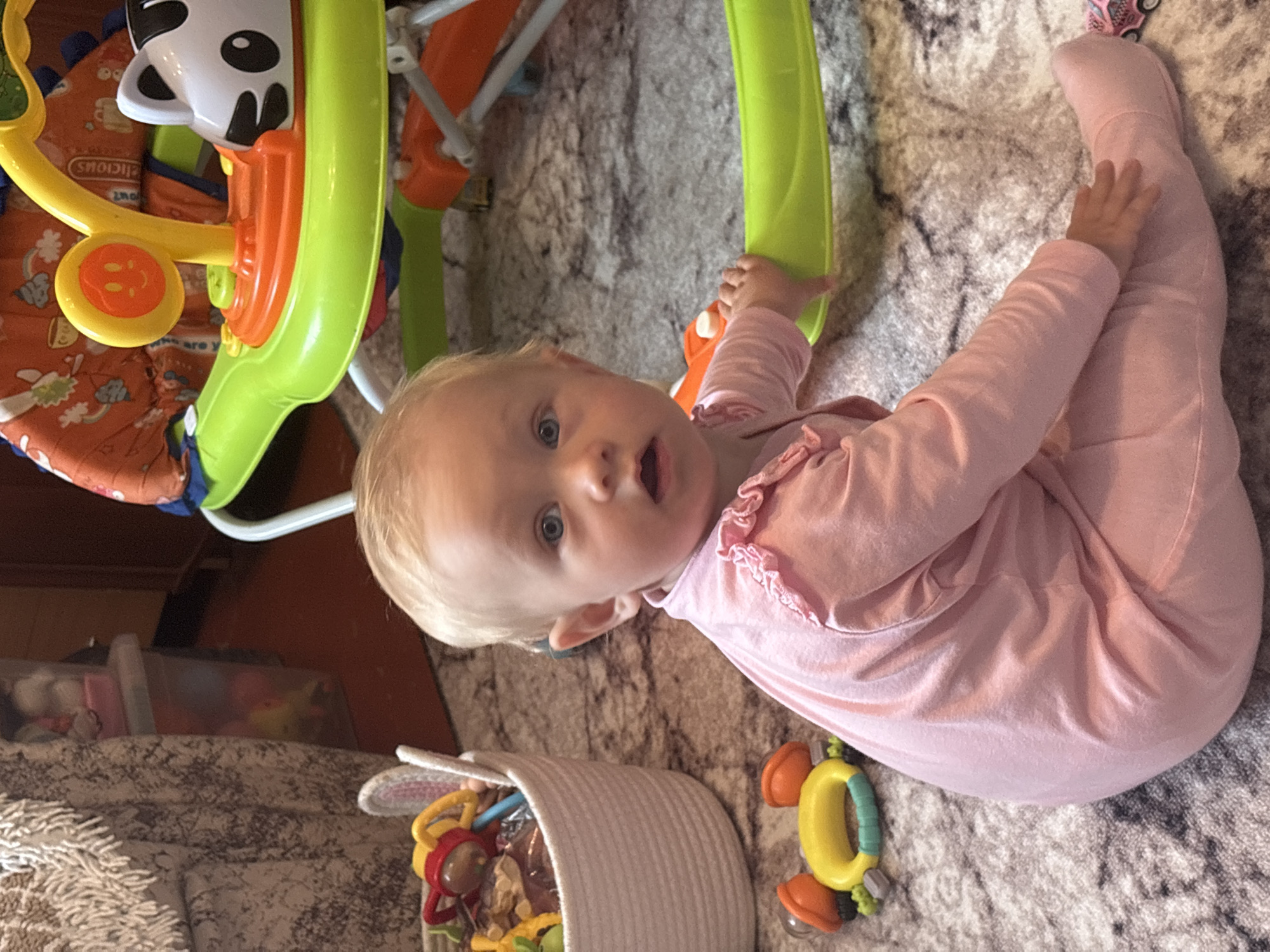 A baby girl is sitting on a rug in what appears to be a home. She is wearing a pink onesie and has blonde hair and blue eyes. She is looking at the camera with a surprised expression. There are toys around her, including a baby walker, a basket of toys, and other smaller toys. The background is blurred but suggests a domestic setting with furniture and other household items. There is no information to indicate this location is in Sovkhoza "Razdol'e", Russia.