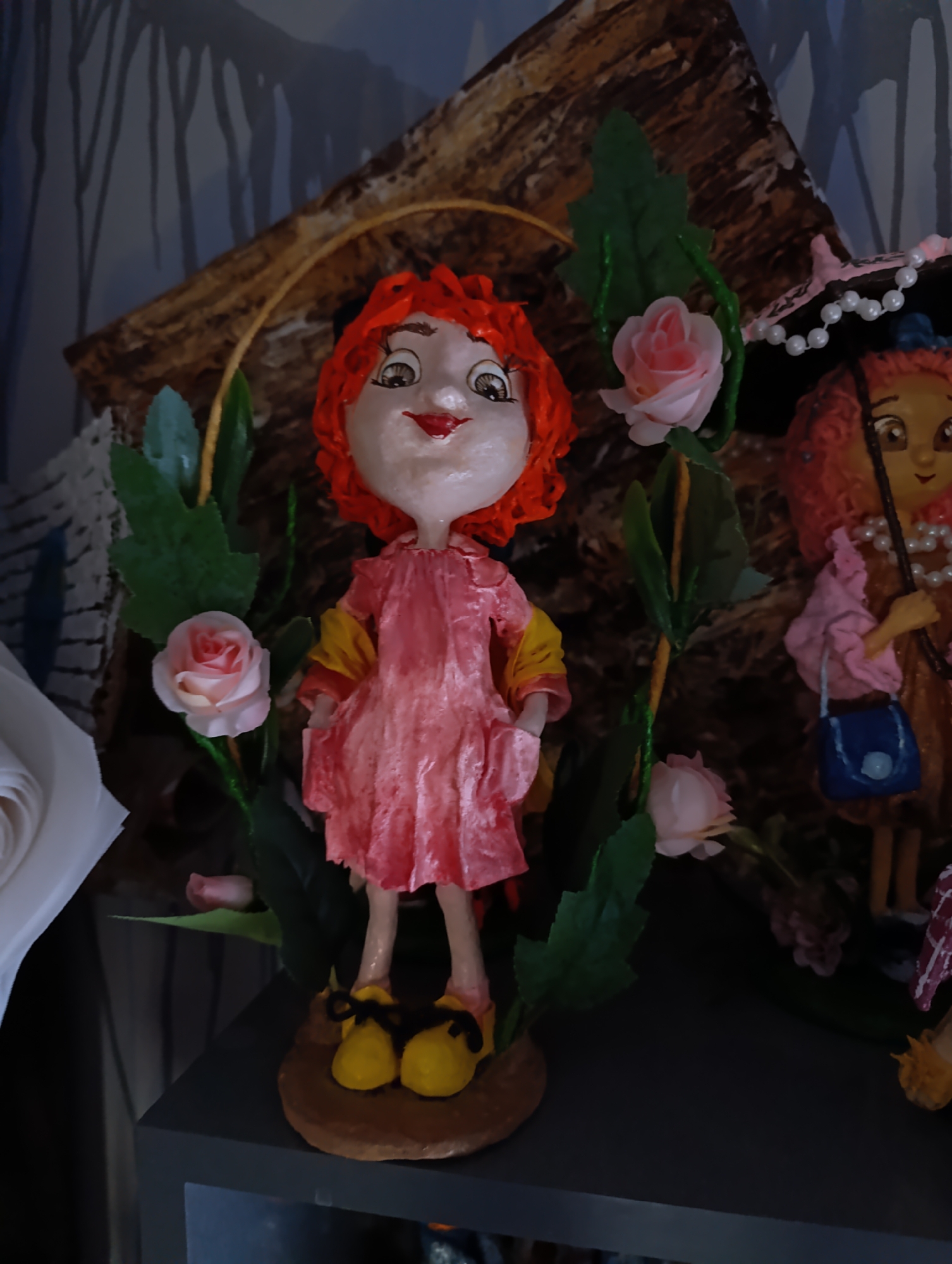 The image shows a close-up of two handmade dolls. The doll in the foreground is a girl with bright red, curly hair, large eyes, and a red mouth. She is wearing a pink dress with yellow accents on the sleeves and yellow shoes. The doll is adorned with green leaves and pink roses. Behind her, a second doll with red hair and a pearl necklace is visible. She is wearing a brown dress and holding a small blue purse. The dolls are displayed on a dark surface, possibly a shelf, with a textured, brown and gray backdrop that resembles a log. The lighting is dim, creating shadows. The scene appears to be an indoor display of crafts. The location in Fergana City, Uzbekistan is not discernible from the image.