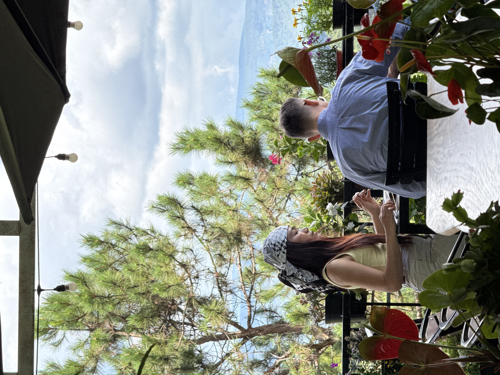Two people sit at a table outdoors. The woman, facing the left of the frame, wears a patterned head covering, a pale yellow top, and light-colored pants. Her hands are near her face. The man, facing right, wears a light blue patterned shirt. The table is white.
The setting includes greenery: trees, bushes, and flowering plants. The sky is visible with clouds. The location is Da Lat, Vietnam.