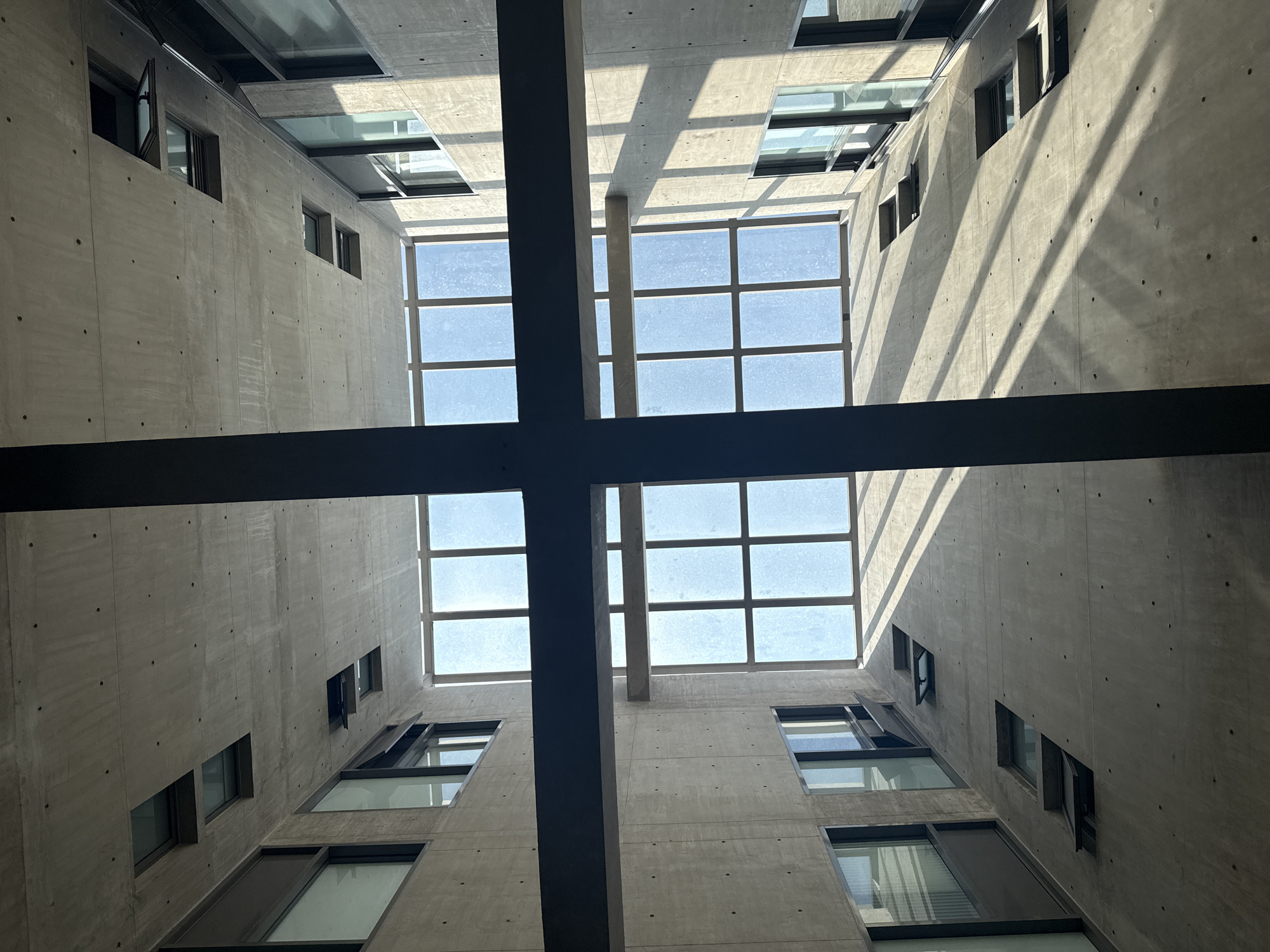 A low-angle shot presents a modern architectural space, likely a courtyard or atrium within a building. The concrete walls rise vertically, punctuated by rectangular windows of varying sizes on multiple floors. The sky is visible through a grid-like skylight system, suggesting a bright, daytime setting. A dark-colored structural support forms a cross in the foreground, adding depth to the composition. The scene conveys a minimalist and industrial aesthetic, indicative of contemporary urban design. The city is Mexico City.