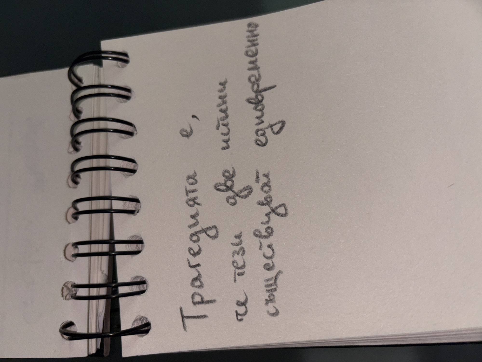 A spiral-bound notepad lies open on a surface, its white pages filled with handwritten text in pencil. The writing is in Cyrillic script and appears to be in Bulgarian. The top line reads "Трагедията е,". The second line begins with "че тези две истини" and the third line continues with "съществуват едновременно". The notepad is partially visible at the top edge of the frame, showing the black metal spiral binding. The background is dark, suggesting an indoor setting, possibly a desk or table. The lighting is low, casting shadows and highlighting the texture of the paper. There are no people or other objects clearly identifiable in the frame. The image does not contain any information that would allow for its location to be determined within Sofia, Bulgaria.