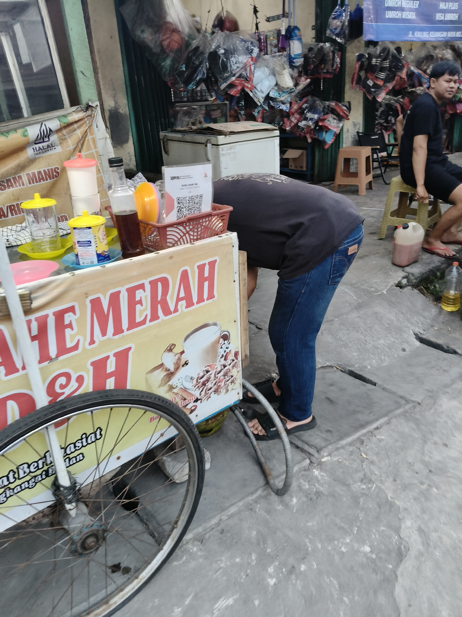 The image depicts an outdoor scene near a street vendor in South Tangerang, Indonesia. A person, dressed in a black t-shirt and blue jeans, is bending over a food cart that displays the text "TAHE MERAH D&H" in large red letters. The cart is adorned with images of a beverage and what appears to be coffee beans or similar items. Various containers, including a bottle of dark liquid, cups, and what looks like a beverage dispenser, are visible on the cart.
To the right of the cart, another person is seated on a light brown plastic stool, also wearing a black t-shirt and shorts. They are looking towards the left of the frame. Behind the seated person, a stack of items, possibly related to travel or religious pilgrimages, are displayed in a cluttered manner, with some items hanging from the ceiling.
In the foreground, the wheel of the food cart is prominently featured. The ground is paved with concrete slabs, showing some wear and tear. A large plastic container with a spigot and another clear plastic bottle containing yellow liquid are present on the ground. The overall environment suggests a bustling street market or a commercial area with multiple vendors. The presence of a "HALAL INDONESIA" logo on a banner attached to the cart further indicates a location within Indonesia. The scene is captured during daylight hours, with natural lighting illuminating the area.