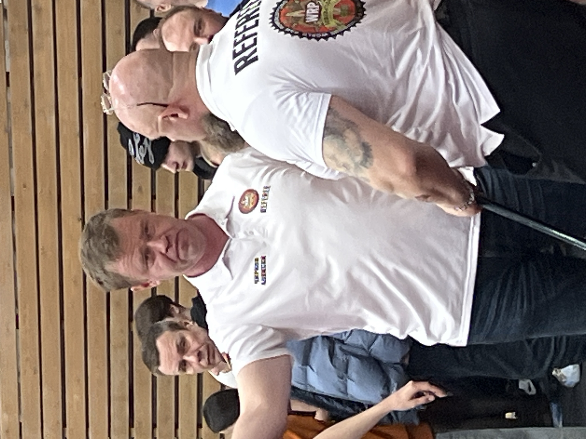 A group of men are gathered indoors, likely at an arm wrestling event. Two men are wearing white polo shirts with "REFEREE" printed on them. One of these men also has a World Armwrestling Professional (WRP) logo on his shirt and an arm tattoo. There are other individuals visible in the background, some partially obscured. The environment includes a wood-paneled wall. The location is not identifiable as Saint Petersburg, Russia based on the visual information provided.