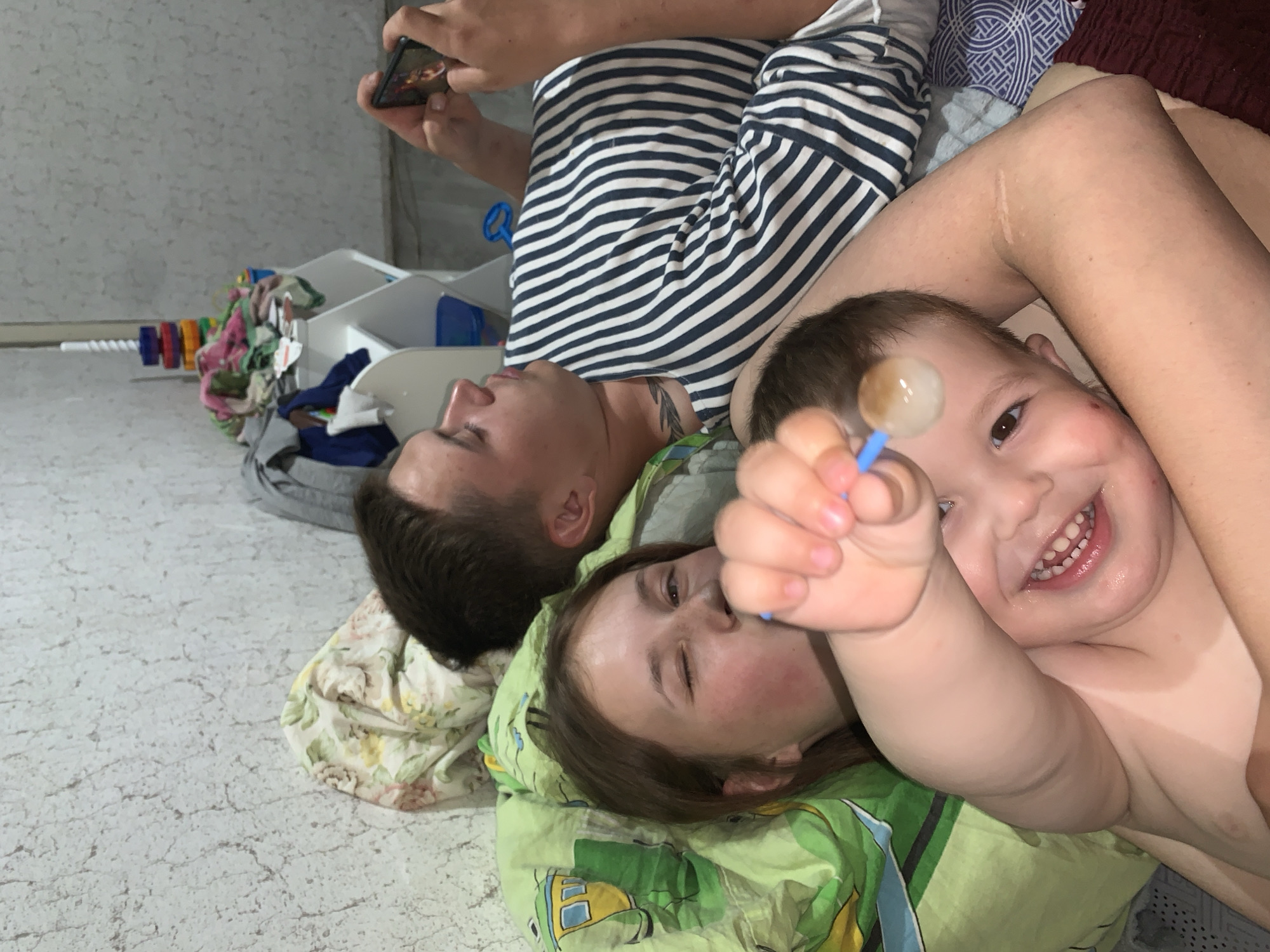 A sideways-oriented, medium shot captures a group of people lying down indoors. The flooring is light-colored with a cracked texture, reminiscent of concrete or stone. In the upper left corner, several white plastic bins are visible, containing assorted items.
A man lies horizontally in the image, wearing a striped shirt. He is holding a smartphone in his hand. Adjacent to him, two young children are present. One of them is holding a lollipop. The lighting suggests an indoor setting, possibly a private residence. Due to limited context, the precise location in Фоки, Russia, cannot be determined.