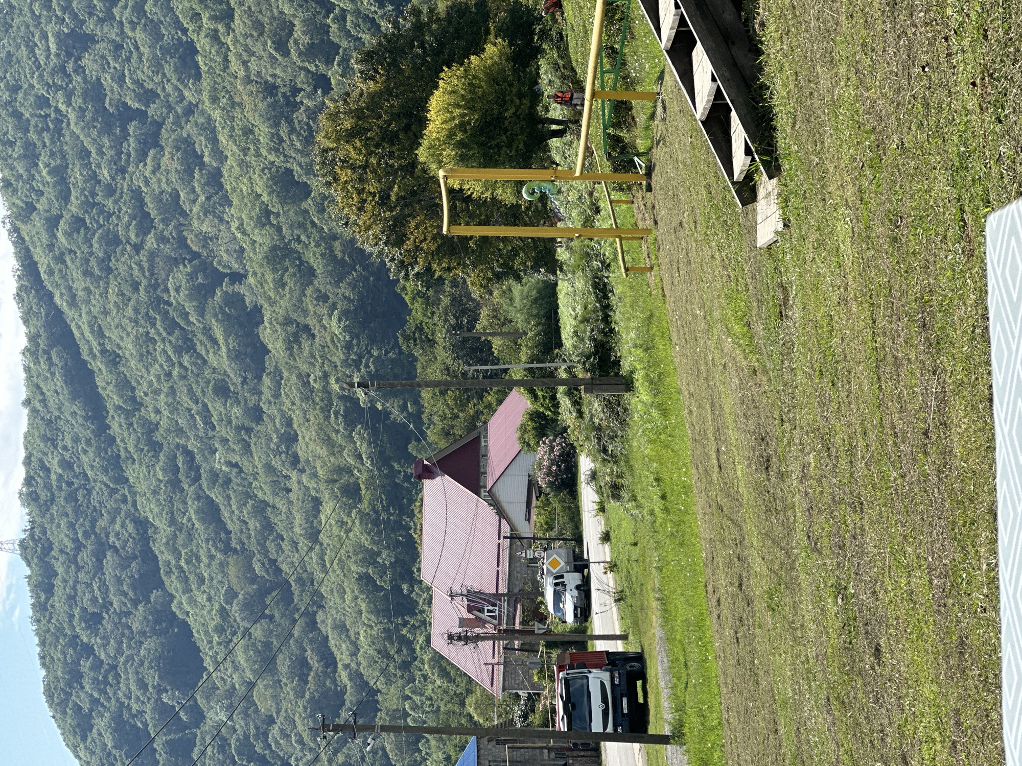 The image depicts a rural landscape in Bolshoy Kichmai, Russia, characterized by a densely wooded mountainside dominating the background. In the mid-ground, a large building with a red metal roof is visible, situated near a paved road. A white van is parked on the roadside in front of the building, and a red truck is parked to the left, facing the road. A yellow metal structure resembling playground equipment or a frame is present in the right mid-ground, with a green inflatable object hanging from it. Wooden stairs lead up from a grassy slope in the foreground. Several utility poles and wires are scattered throughout the scene. The overall environment is green and appears to be a sunny day.