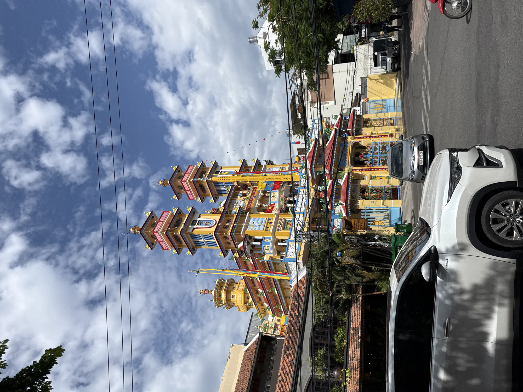 This image captures a street view in Nha Trang, Vietnam, dominated by a vibrant, multi-tiered Buddhist pagoda with yellow and red accents. The pagoda features numerous towers, decorative elements, and colorful flags. In the foreground, a white SUV is parked on the street, its shadow cast across the vehicle. Across the street, several other cars, including a silver sedan and a silver van, are visible. People are seen walking on the sidewalk and near the entrance of the pagoda. The environment includes trees, power lines crisscrossing the sky, and a blue sky with scattered clouds. The architecture of the pagoda suggests it is a significant religious and cultural landmark.