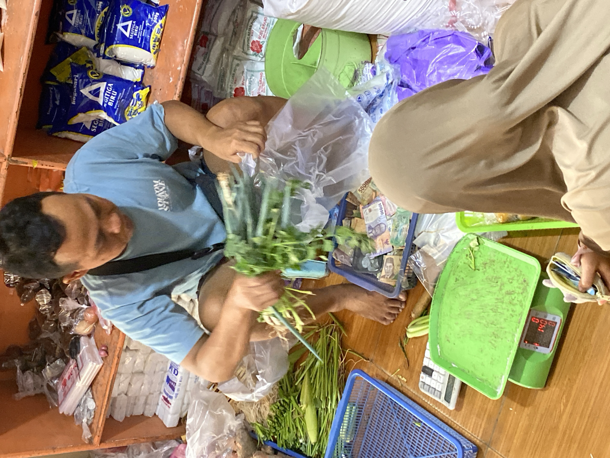 In a market stall in Kasihan, Indonesia, a man wearing a light blue t-shirt is shown crouched down, sorting through a bunch of green vegetables, possibly herbs or leafy greens. He holds the vegetables in his right hand, with his left hand resting on a plastic bag. In front of him, a green weighing scale displays the number "140", suggesting he is weighing the produce. Next to the scale is a calculator. A portion of a person wearing a beige hijab is visible in the foreground, facing away from the camera. The stall is filled with various goods, including bags of flour or other dry goods stacked on shelves behind the man. Baskets containing more vegetables and some Indonesian Rupiah banknotes are also present in the scene. The floor appears to be made of wood.