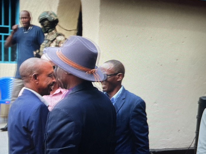 The image depicts a group of four men standing in front of a white wall in a building in Keshero, Democratic Republic of the Congo. One of the men is wearing a blue hat and a dark suit jacket. Another man is wearing a light blue shirt and a dark suit jacket. The two men are looking at each other and smiling. There are two additional men in the background. The man on the left is wearing a dark blue shirt and the man on the right is wearing a light blue shirt. A statue of a soldier wearing a helmet and camouflage is positioned in the background. A light blue plastic chair is positioned in the background. The scene is shot in a close-up perspective.
