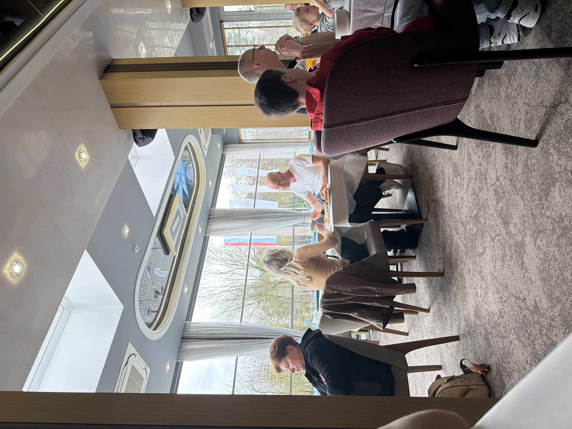 The image shows an interior scene, likely a restaurant or dining area, with several individuals seated around tables. The location is possibly Vidin, Bulgaria, as suggested by the flags visible through the windows, but this is not definitively confirmed. There are multiple people in the image: A person in a brown jacket is seated at a table, appearing to be deep in thought or looking down. Adjacent to them, a woman with blonde hair is seated at the table, possibly interacting with someone. Across the table, a man in a white shirt appears to be handling food. In the background, there's another table where a person in a red shirt is seated, and next to them, a bald man is engaged in conversation with a young boy. The environment is well-lit with large windows providing a view of the outside, where trees are visible. The decor of the dining area includes an artistic design on the ceiling, and the room is furnished with modern chairs and tables. A purse is on the floor. The overall impression is a gathering of people, possibly a family or group of friends, enjoying a meal together.